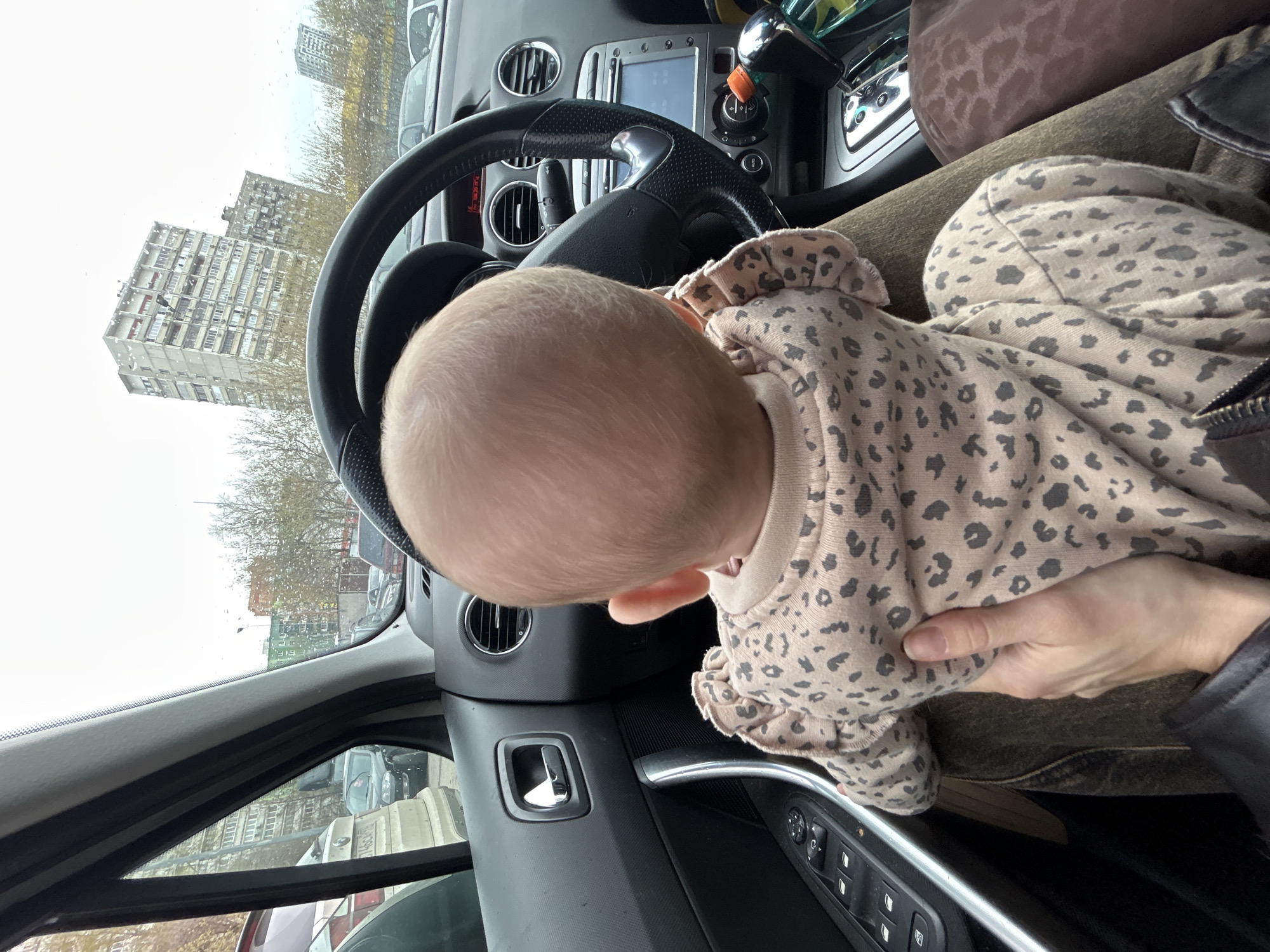 A baby in a spotted onesie is held in the lap of someone sitting in the driver's seat of a car. The car is parked, with a view of a building in Moscow, Russia, visible through the windshield.