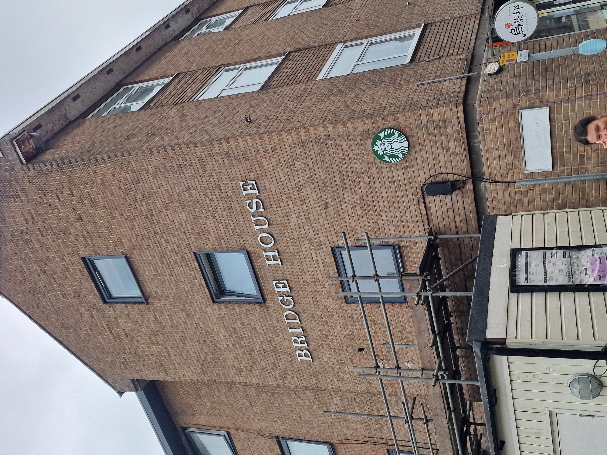 The image depicts a portion of a multi-story brick building labeled "BRIDGE HOUSE" with a Starbucks logo prominently displayed. The building has several windows. Scaffolding is present in front of the building, partially obstructing the view of the entrance. A person is partially visible on the right side of the image. A street sign with Chinese characters is also present. The scene is located in the city of Durham, United Kingdom.