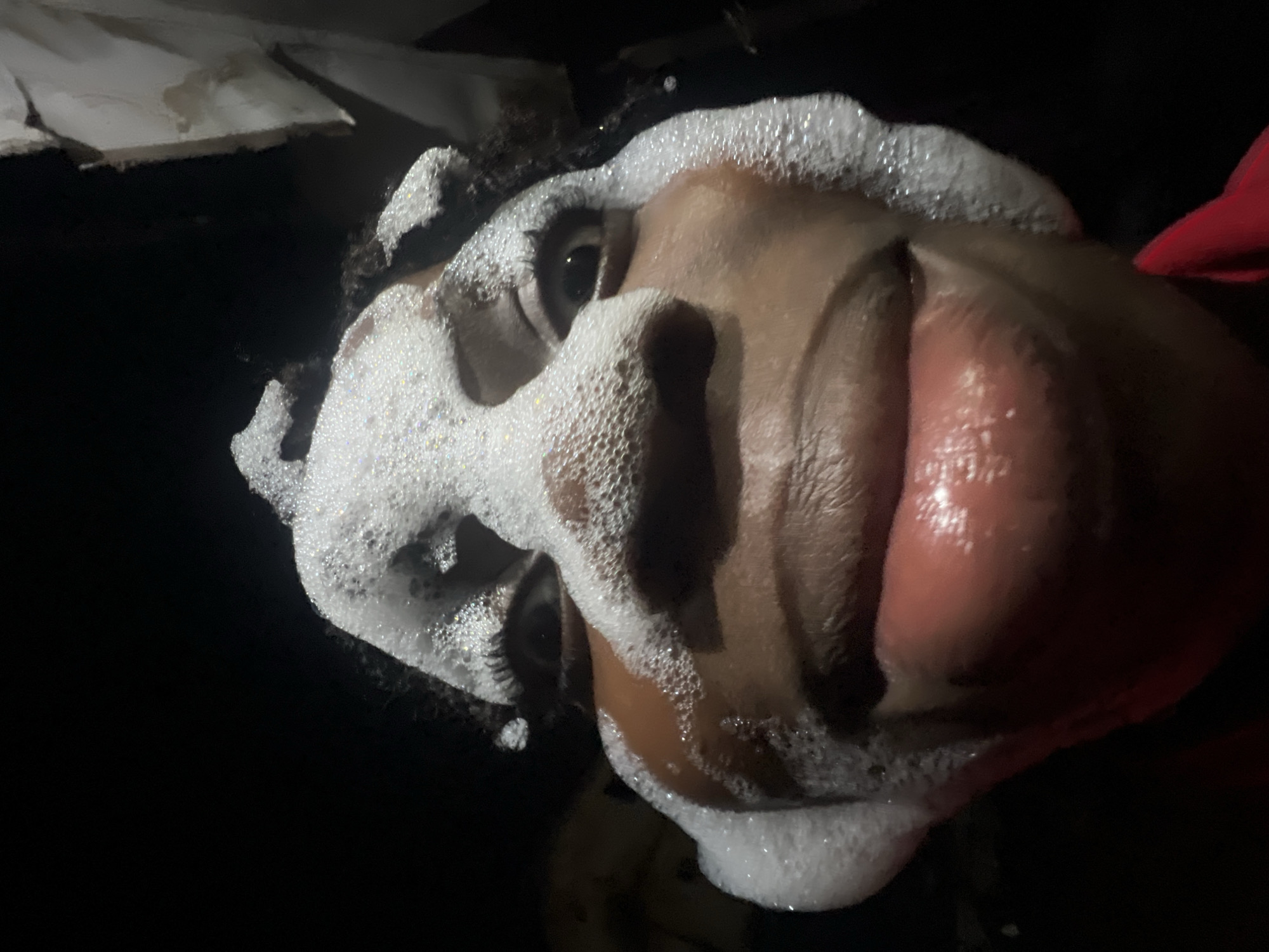 A close-up, low-angle, front-facing shot captures a person's face covered in white foam, seemingly from a bath or shower. The foam obscures much of their features, but their eyes are visible, wide open and looking directly at the camera. The individual's lips are also visible, slightly parted and glistening, with some foam on the side of their face near the mouth. The background is dark and indistinct, suggesting an indoor setting at night. Some textured, off-white material is visible in the upper right corner, possibly a wall or ceiling. The overall impression is of a playful or experimental moment captured in a private space in Nkpor, Nigeria.