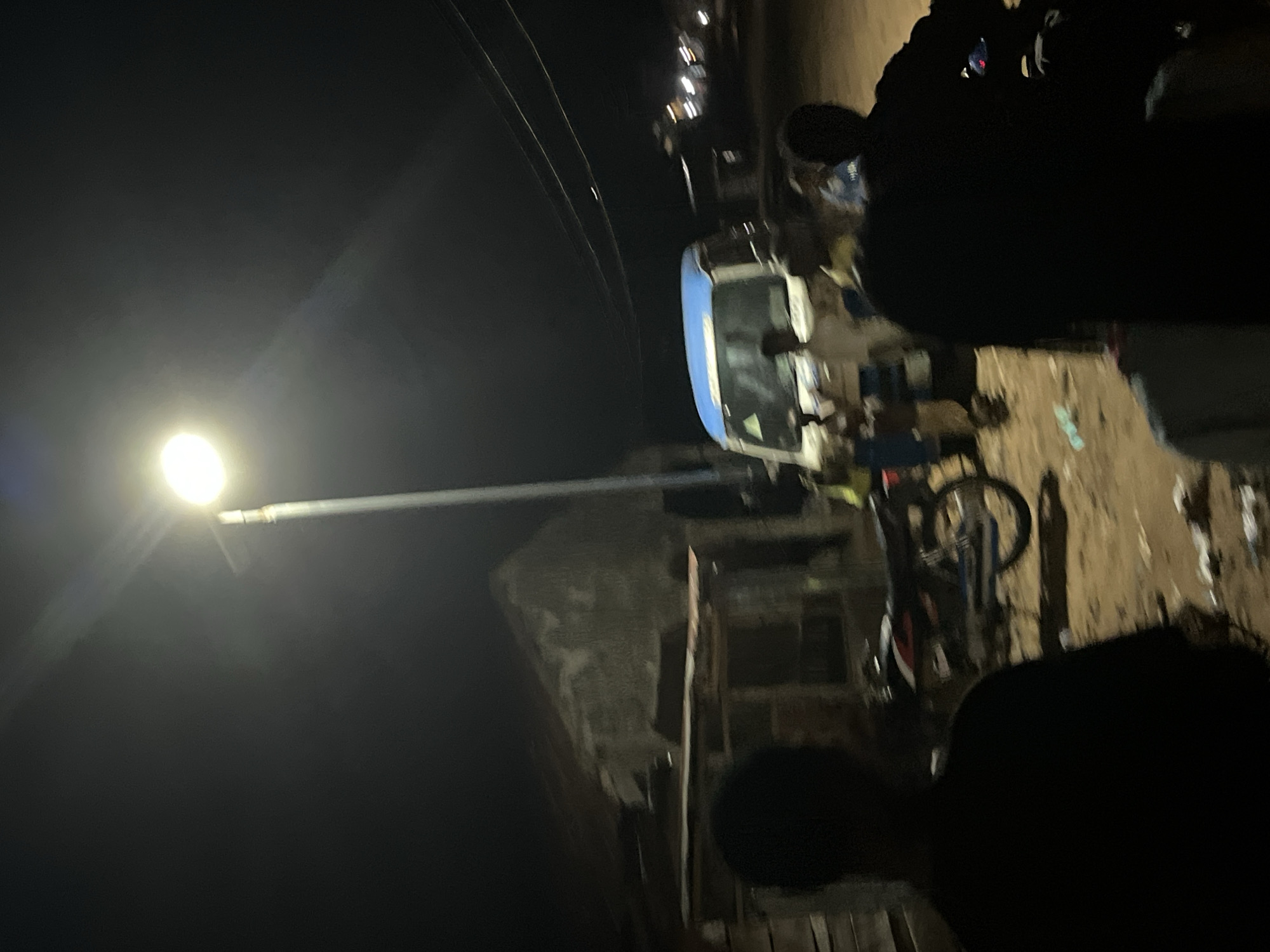 The image captures an outdoor night scene illuminated primarily by a single, bright street light positioned on the left side of the frame. The time of day is dark night, with deep shadows obscuring much of the environment and figures.
In the midground, a light-colored vehicle, appearing to be a white van or small bus with blue accents, is visible to the right of the street light. A bicycle is parked or leaning near the vehicle. The ground beneath appears unpaved, uneven, and possibly dusty or muddy.
Several individuals are present, mostly rendered as dark silhouettes against the ambient light. One person's head and shoulders are prominent in the bottom right foreground. Other figures are visible further back, near the vehicle and surrounding what appear to be informal structures or market stalls. Overhead, thin lines, likely power or communication wires, traverse the dark sky. The scene suggests a bustling area, possibly a street or communal space, at night. No specific activity is clearly discernible beyond the presence of people and objects under artificial light. There is no visible text in the image.