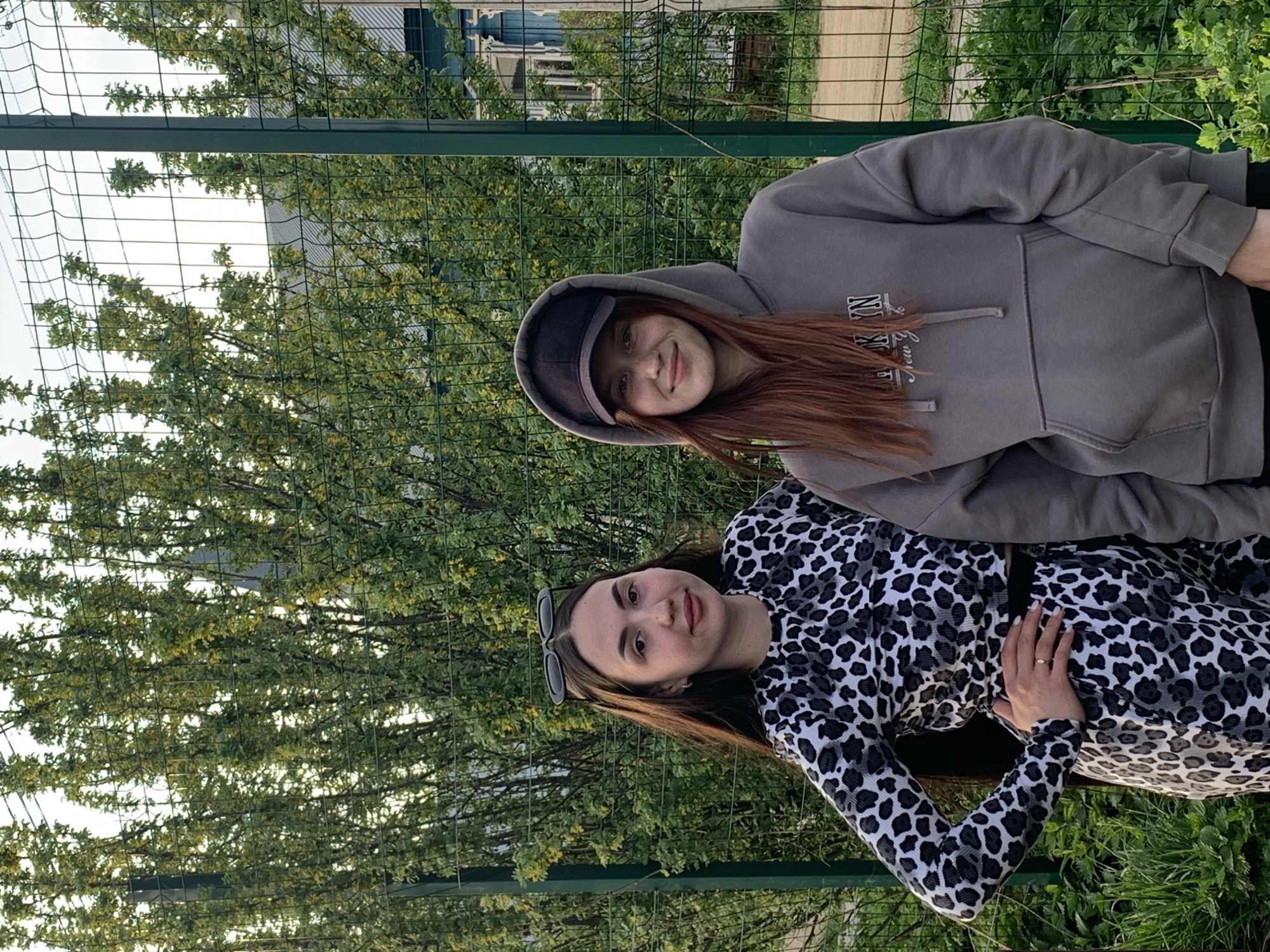 Two women are posing for a photo outdoors in Фоки, Russia. The woman on the right wears a gray hooded sweatshirt and a dark cap. Her red-brown hair is visible beneath the cap and around her shoulders. The woman on the left wears a long-sleeved, spotted animal print top. Sunglasses are perched on top of her head, and her hair is long and brown. Her left hand rests on her body. The background features a green chain-link fence, trees, and buildings.