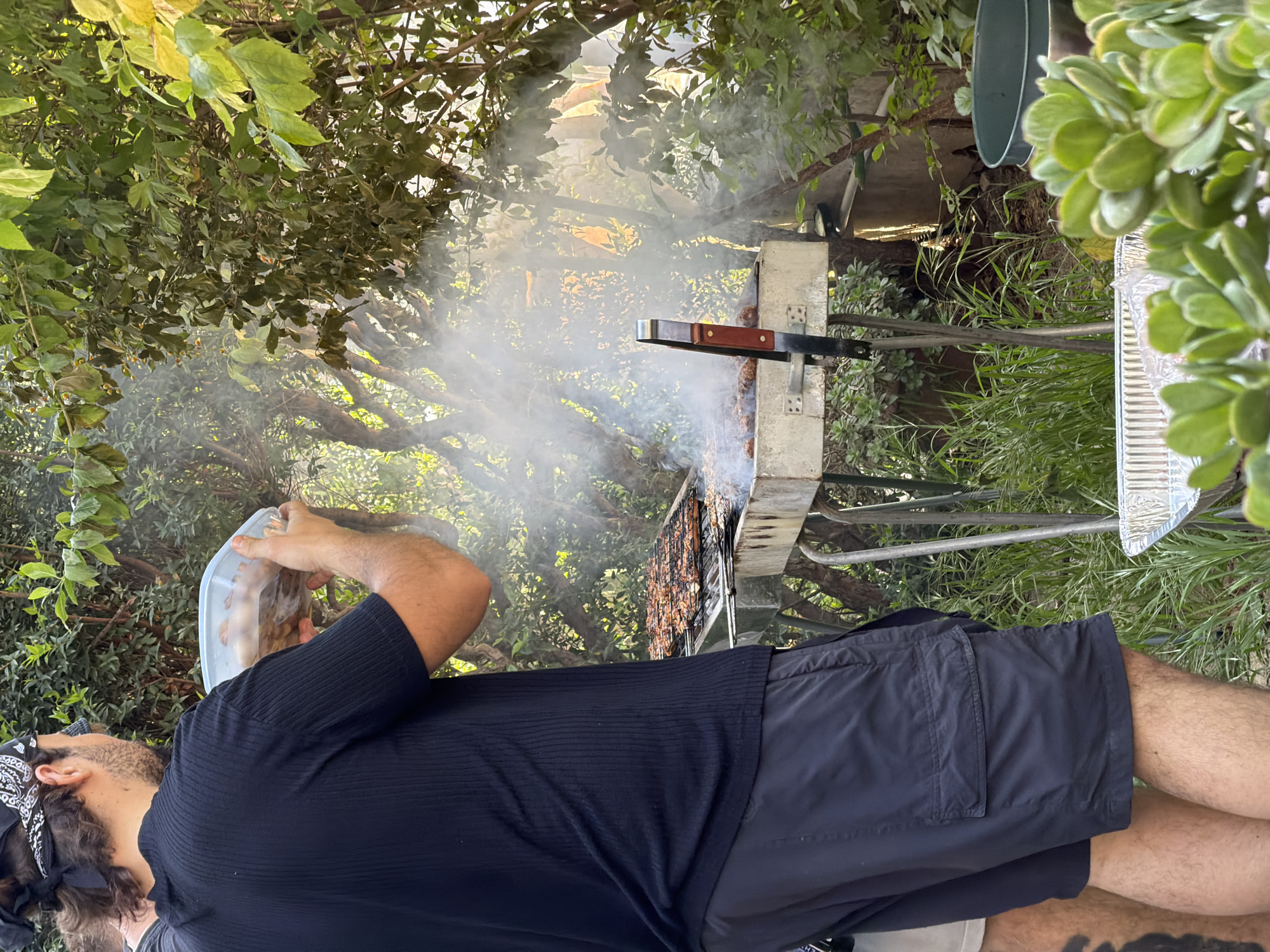 A man with a bandana and dark hair, wearing a dark short-sleeved shirt and dark shorts, is grilling food outdoors, possibly in Kiryat Motzkin, Israel. He is holding a container with food in his left hand, while smoke rises from the grill. The grill is set against a backdrop of trees and greenery. A foil tray and cooking utensils are visible near the grill.