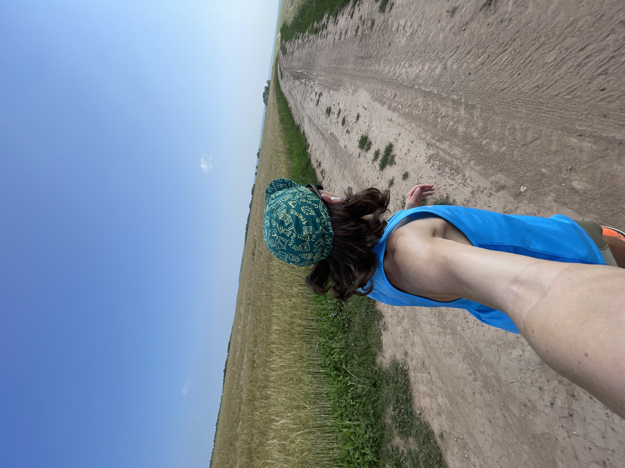 A first-person perspective shot captures a person from the chest up, facing away from the camera, as they walk along a dusty dirt road. The individual is wearing a bright blue tank top and a green bandana with white text and graphics. Their arm is extended forward, suggesting they are holding a camera or phone to capture the scene. The road stretches into the distance, winding through a vast, golden field of ripe wheat under a clear blue sky. Green foliage lines the left side of the road, contrasting with the yellow field. In the far distance, a faint treeline and a few structures can be seen, hinting at a rural setting, possibly the outskirts of Maksimovo, Russia.