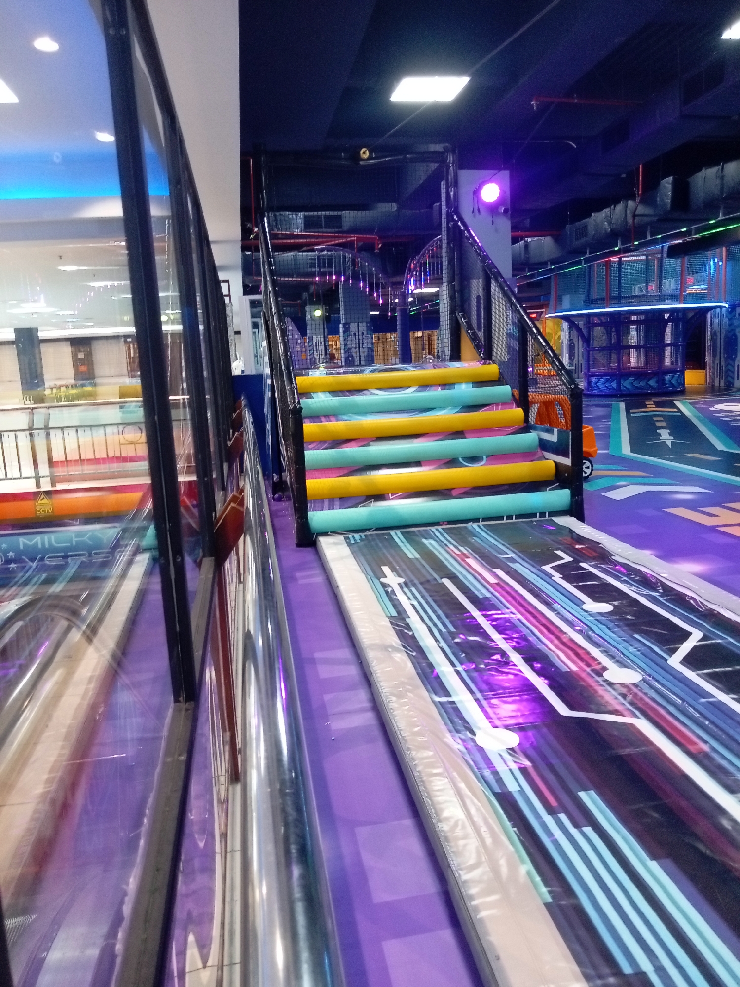 This image depicts an indoor amusement park in Batam, Indonesia, likely located within a shopping mall. The scene features a colorful staircase with alternating yellow and teal cushioned steps. To the right of the stairs, a futuristic, brightly lit floor graphic is visible, with purple and blue lines creating a dynamic pattern. In the background, a netted play structure with blue and red accents is apparent. The overall environment suggests a lively and engaging space for children and families.