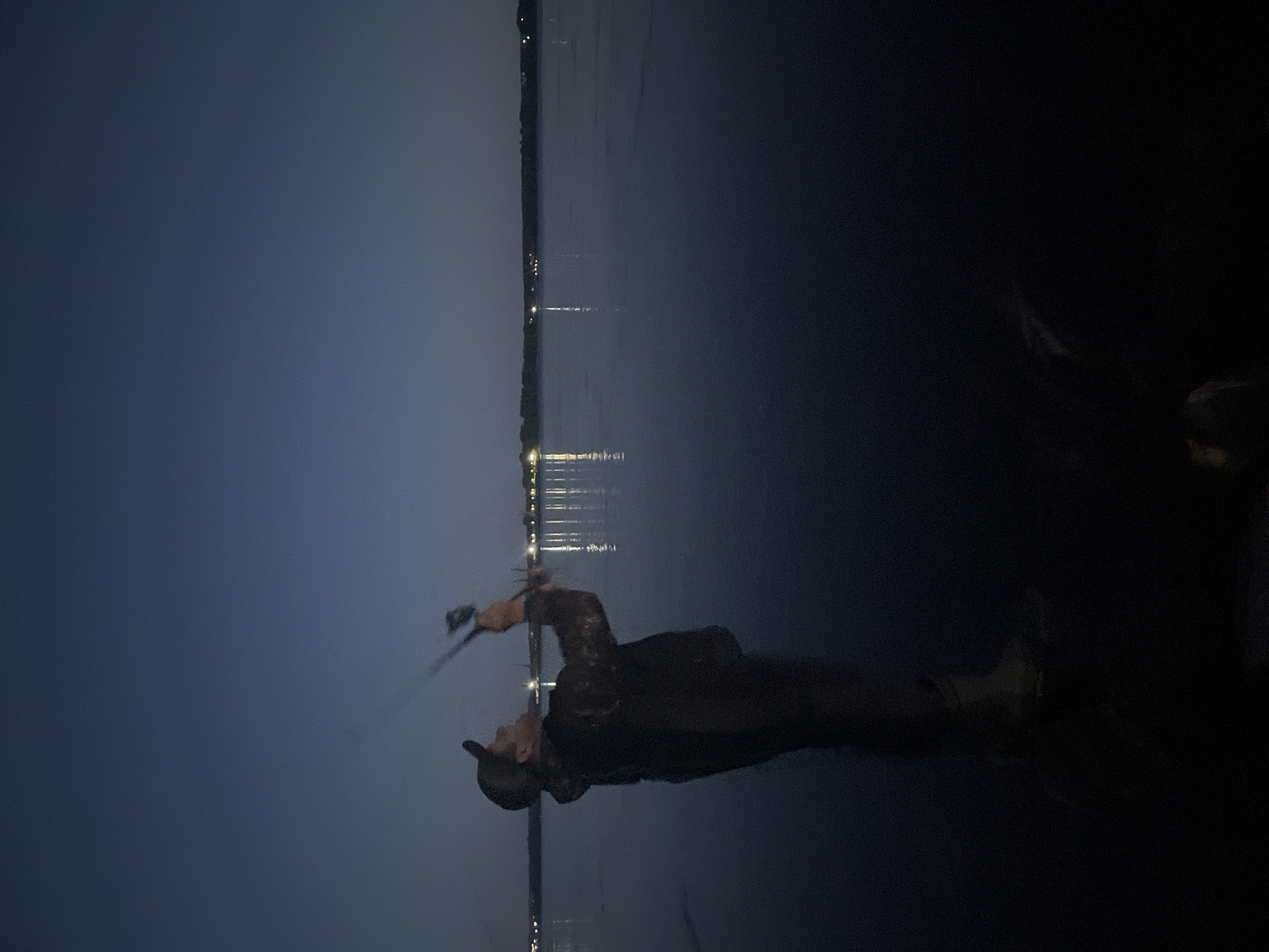 A lone fisherman, wearing a camouflage jacket and a cap, stands on the shore of a body of water at dusk. He is casting a fishing rod, with the line extending upwards into the darkening sky. The water is calm, with a faint reflection of distant lights on the horizon. The background shows a treeline and a few scattered buildings, indicating a populated area. The overall atmosphere is peaceful and serene, with the natural beauty of the landscape illuminated by the fading light. This scene is located in Chaikovsky, Russia.