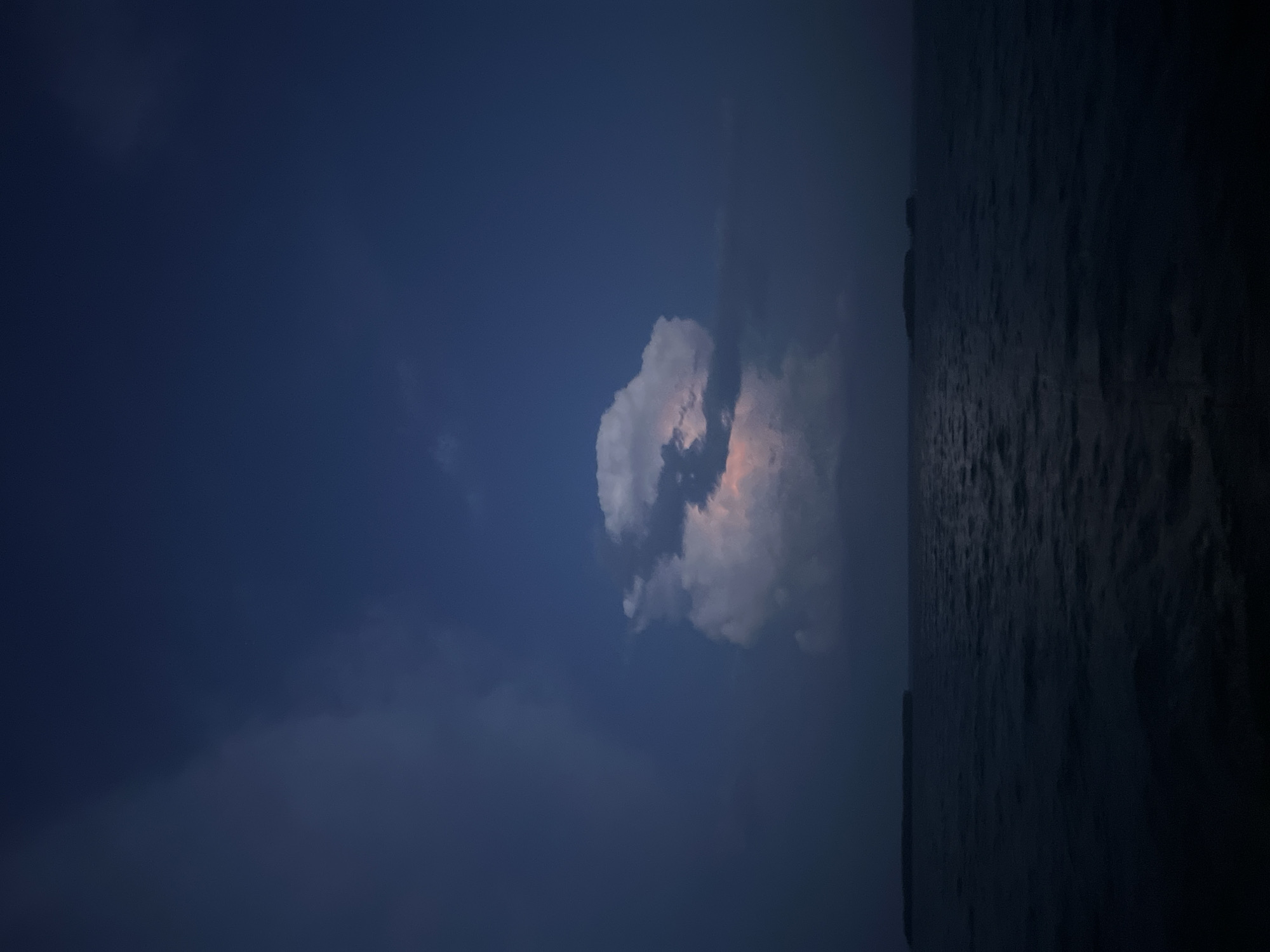 A dark blue sky with scattered clouds is visible above a dark body of water. A single, brightly lit cumulus cloud is in the center of the frame, appearing to glow from within with pink and white light. Two distant, dark land masses are visible on the horizon, one on the left and one on the right. The water has small ripples.