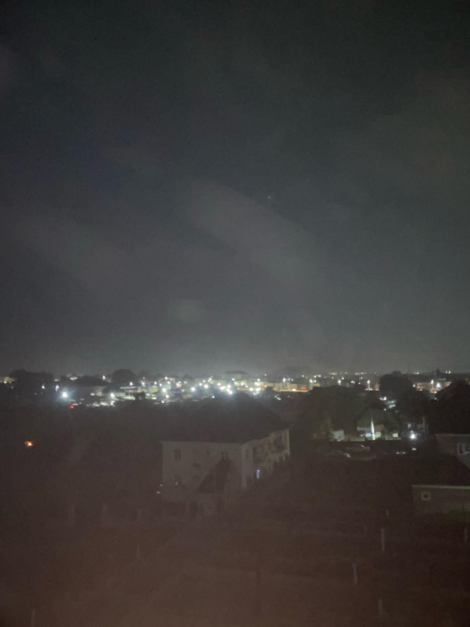 The image shows an aerial view of a residential area at night in Abuja, Nigeria. The sky is dark with some wispy clouds, and the city lights create a hazy glow below. In the foreground, several multi-story buildings are visible, with some lights on inside. The architecture appears to be modern, with a mix of flat roofs and some pitched roofs. The overall impression is of a quiet, suburban neighborhood. The image is slightly out of focus, giving it a dreamy or atmospheric quality.