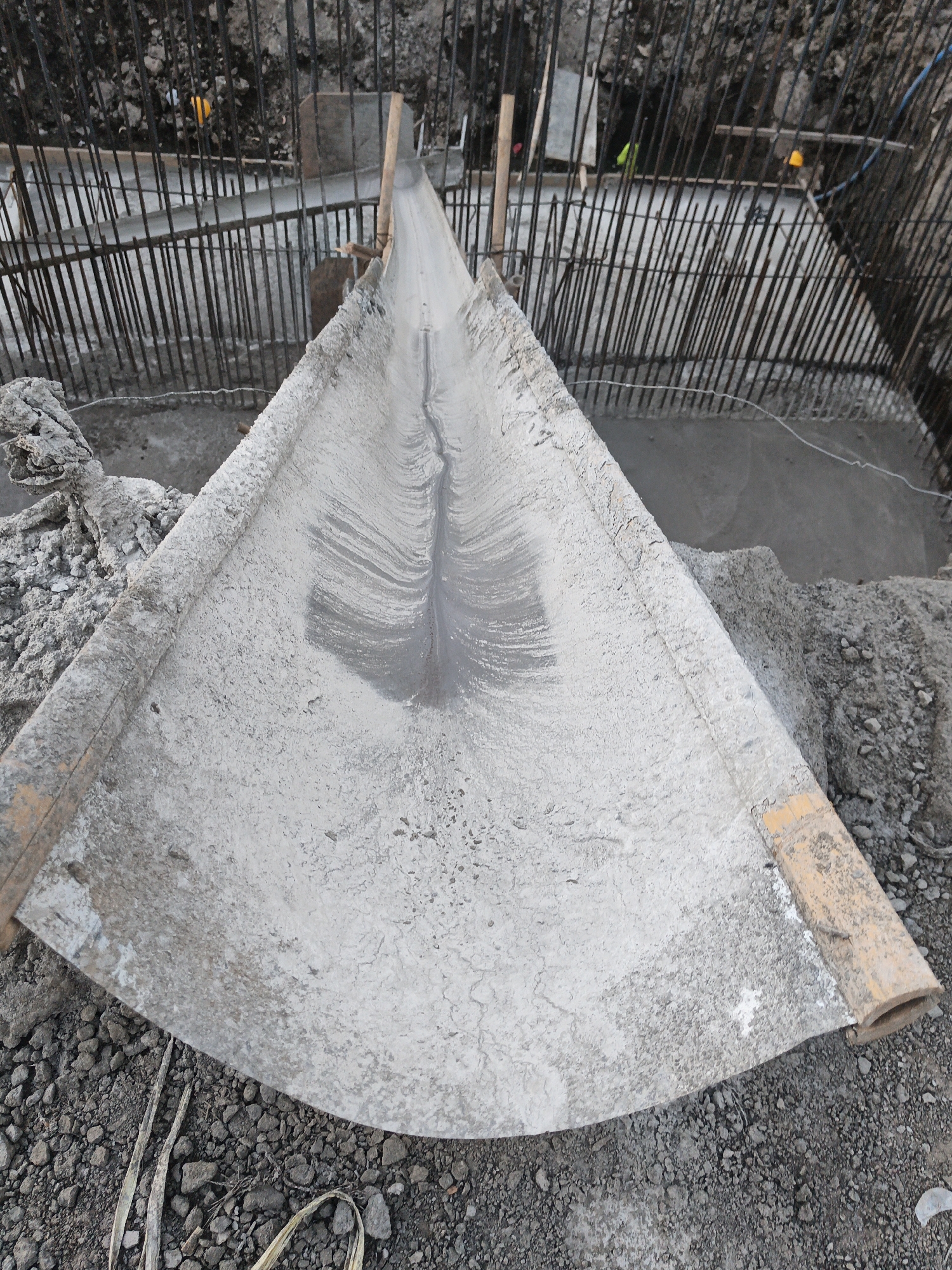 A construction site in Planggu, Indonesia, features a large, trapezoidal concrete chute, partially filled with wet concrete. The chute is positioned above a reinforced concrete structure, with vertical rebar rods visible throughout the site. Several piles of aggregate and sand are scattered around the chute. Two yellow hard hats are visible in the background, and a person in a blue shirt is partially visible, possibly overseeing the concrete pour. The scene suggests ongoing construction work on a foundation or structural element.
