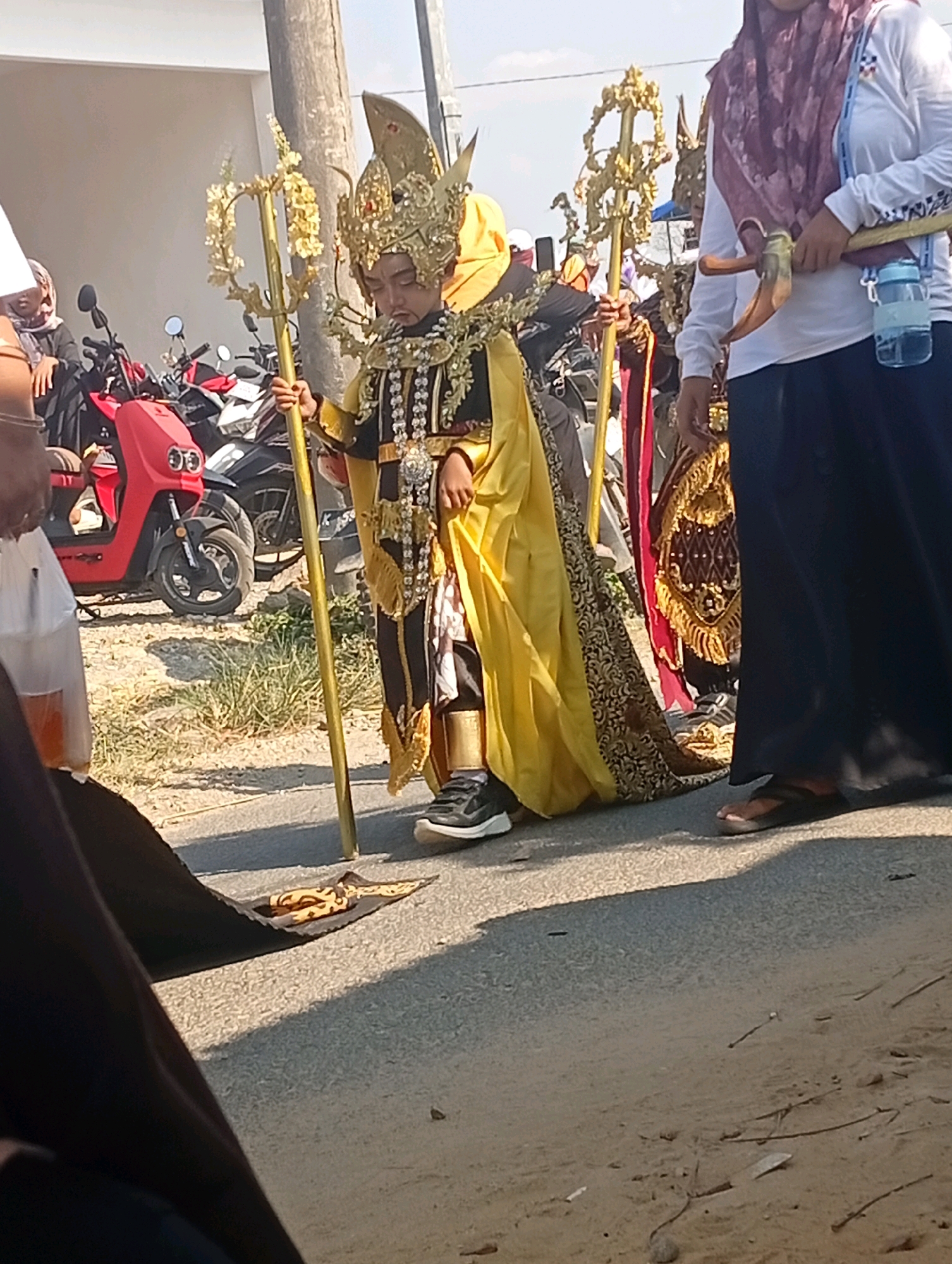 In a street scene in Nglojo, Indonesia, a young boy dressed in an elaborate, golden costume adorned with intricate black and gold patterns, and a matching golden headdress, stands holding a decorative golden staff. He is positioned on asphalt next to a grassy area. To the left of the boy, a person wearing a white hijab and dark clothing is partially visible, holding a clear plastic bag containing a beverage. Behind them, several motorcycles are parked. To the right of the boy, a woman wearing a burgundy hijab and white long-sleeved shirt is visible. She is carrying a tool resembling a pickaxe and a blue water bottle. The ground around them is a mixture of asphalt and sandy dirt with scattered debris. The sky is clear and bright.