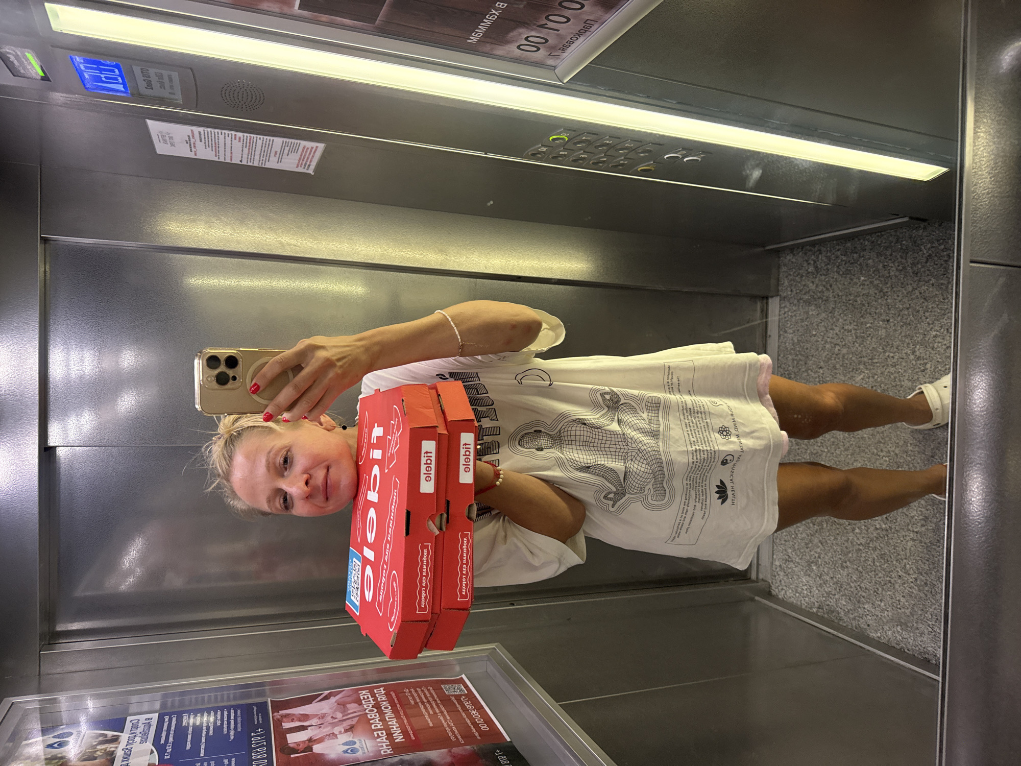 A woman stands in an elevator holding two red pizza boxes. She is wearing a white t-shirt with a graphic of a meditating figure and a pink garment underneath. She has blonde hair pulled up in a bun and is taking a selfie with her phone. The elevator has mirrored walls and a textured floor. A digital display on the elevator wall shows the number 12 and a green illuminated button. There are posters on the wall to the left of the woman, one with text in Russian.