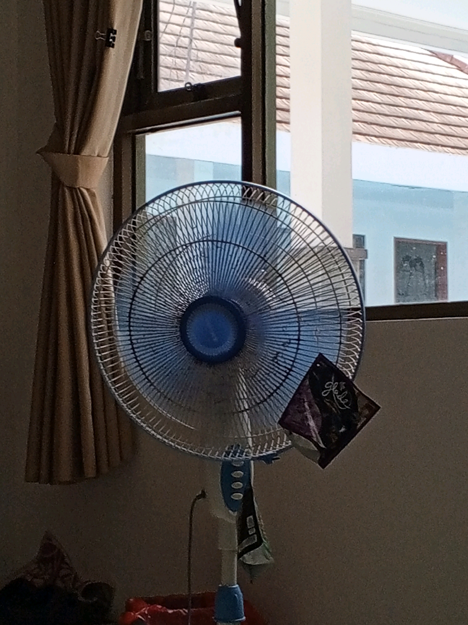 The image depicts a standing fan positioned in front of a window, with a curtain to its left. A small, square object, possibly a sachet or card with text that appears to read "Glace," is attached to the fan's grille. The background reveals a window with blinds and a view of a tiled roof and a light blue building, suggesting an indoor setting in a residential or commercial space. The overall lighting is dim, indicating it could be daytime with limited natural light or evening. The fan is the central subject, with its blue motor housing and silver grille clearly visible. The presence of a sachet suggests it might be an air freshener or a similar item. The scene appears to be a common household or office environment, possibly in Denpasar, Indonesia, given the architectural cues and the typical use of such fans in warmer climates.