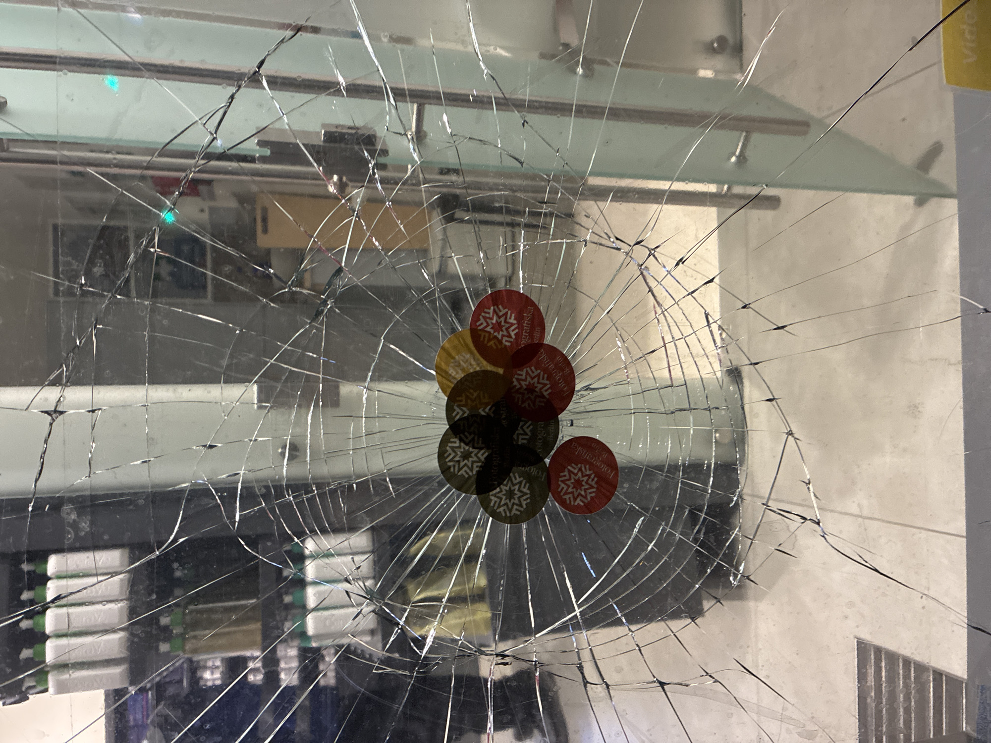 The image shows a close-up of a cracked glass pane, likely a storefront window. Through the shattered glass, a display of products, possibly toiletries or cleaning supplies, can be seen on shelves. Several circular stickers, some red, green, and gold, are affixed to the glass, each featuring a snowflake-like design and text that reads "Fotografiska Berlin." The background suggests an indoor setting, possibly a retail establishment or gallery, with a door frame and handle visible to the right. The overall impression is one of damage and vandalism to a commercial space. There are no people visible in the image. The location is identified as Berlin, Germany, based on the text on the stickers.