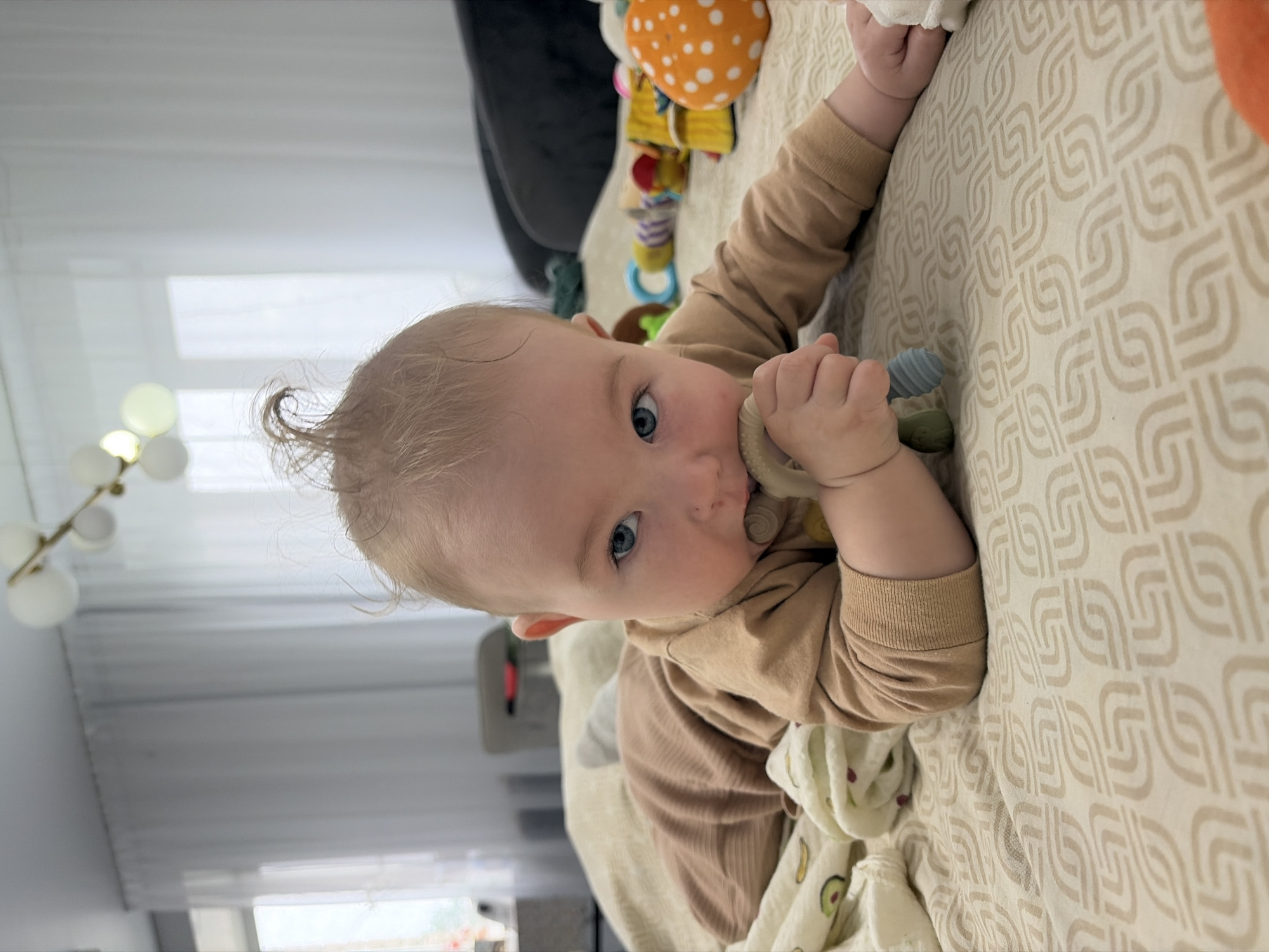 A close-up, eye-level shot captures a baby lying prone on a patterned beige blanket indoors. The baby, with blonde hair styled in a peculiar tuft, has blue eyes and is chewing on a light-colored teething toy. The baby wears a light brown long-sleeved shirt and light brown ribbed pants. To the left of the baby, a white swaddle with fruit patterns (avocado and bananas) is visible. The background features sheer white curtains behind a window, a modern chandelier with multiple globe lights, and glimpses of furniture. Several colorful baby toys, including a mushroom-shaped toy with orange and white polka dots, are scattered around the baby on the blanket. The overall environment suggests a domestic setting, likely a child's play area or bedroom. The image does not provide specific geographical information to pinpoint its location within Chaikovsky, Russia.