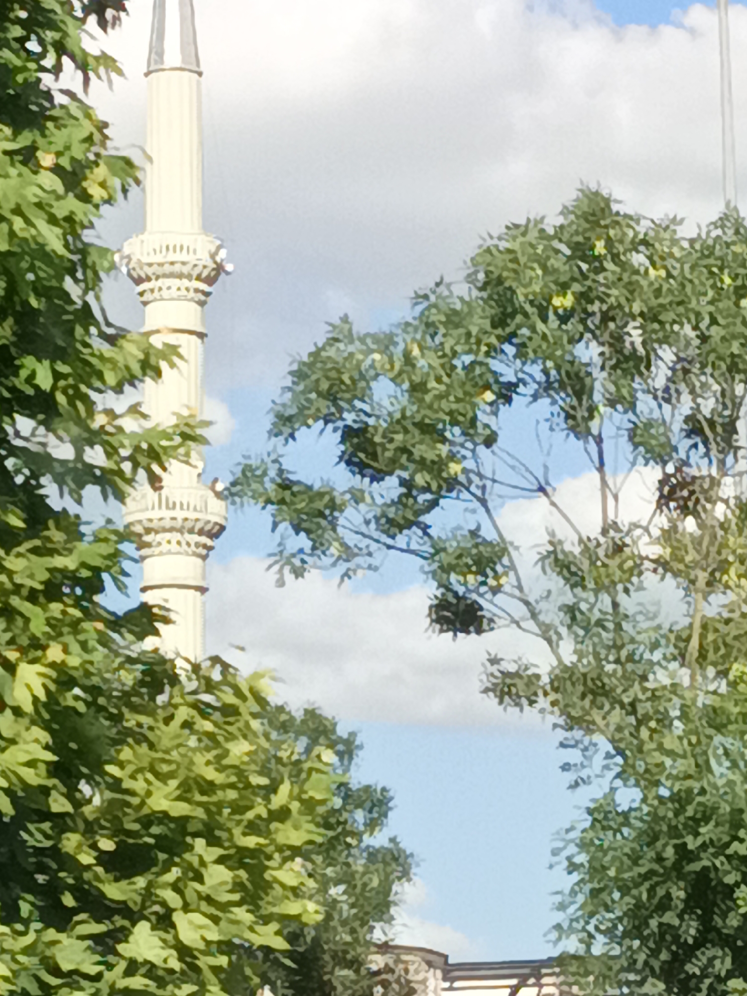 This media file depicts a close-up view of a minaret of a mosque, partially obscured by lush green trees. The minaret is tall and slender, with fluted architectural details and decorative balconies, rendered in a light, creamy color. Above the balconies, the minaret tapers to a pointed spire.
The trees frame the minaret, with dense foliage in shades of green, suggesting it is either spring or summer. The leaves are varied in size and shape, creating a textured canopy. Sunlight filters through the branches, dappling the scene.
The background shows a bright blue sky with scattered white clouds, indicating fair weather. The lighting suggests it is daytime, likely late morning or afternoon. The overall impression is one of peacefulness and natural beauty surrounding a religious structure.
In the lower part of the image, hints of buildings with arched elements and tiling can be seen beneath the trees, suggesting an urban or suburban setting. The scene appears to be located in Mamak, Turkey, as indicated by the location context. No people are clearly visible, nor is there any text present in the image. The focus is on the architectural element of the minaret and the surrounding nature.
