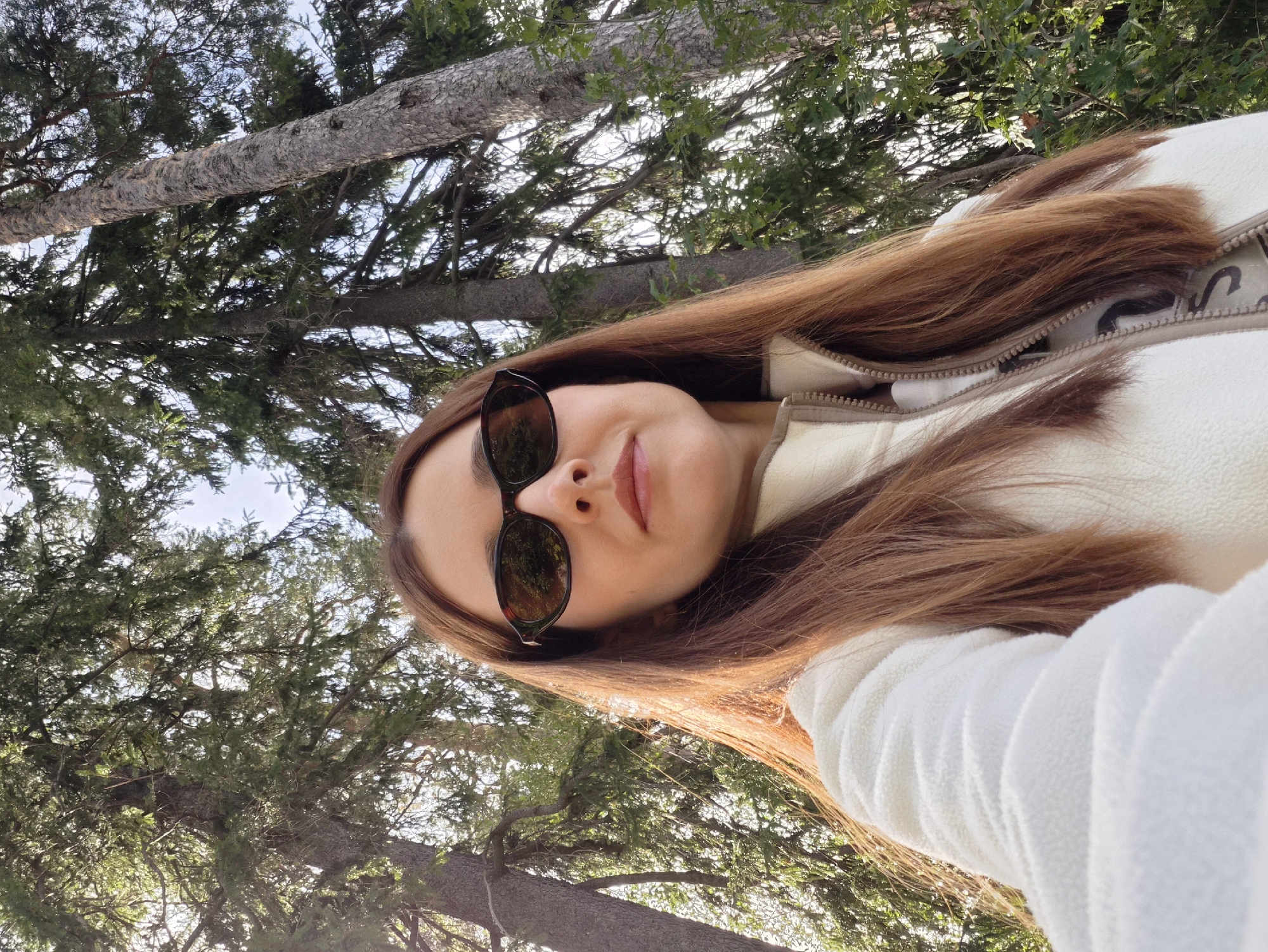 A low-angle, medium shot captures a woman with long, auburn hair and sunglasses, her face partially obscured by them. She is wearing a cream-colored fleece jacket zipped up to her neck. She is outdoors, surrounded by tall evergreen trees. The sunlight filters through the branches, casting a warm glow on her face and hair. The image is devoid of any specific Vyborg landmarks, suggesting a natural setting that could be found within or near the city. There are no other people or animals visible.