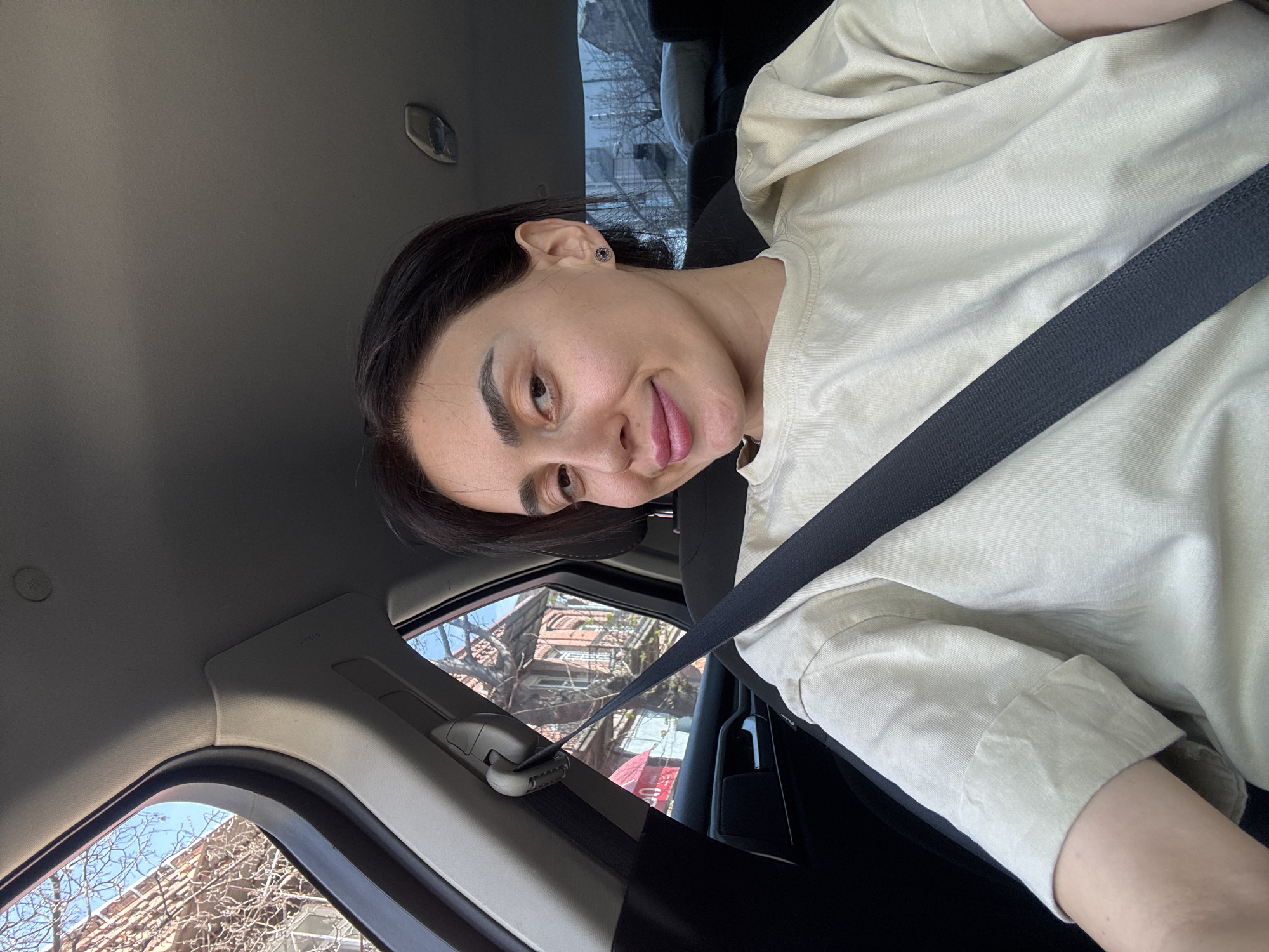A woman with dark hair and a light complexion is pictured from a slightly low angle inside a car, wearing a seatbelt. She is wearing a light beige shirt. Her tongue is slightly extended. A building with bare trees in front of it is visible through the car window. The location is Tbilisi, Georgia.