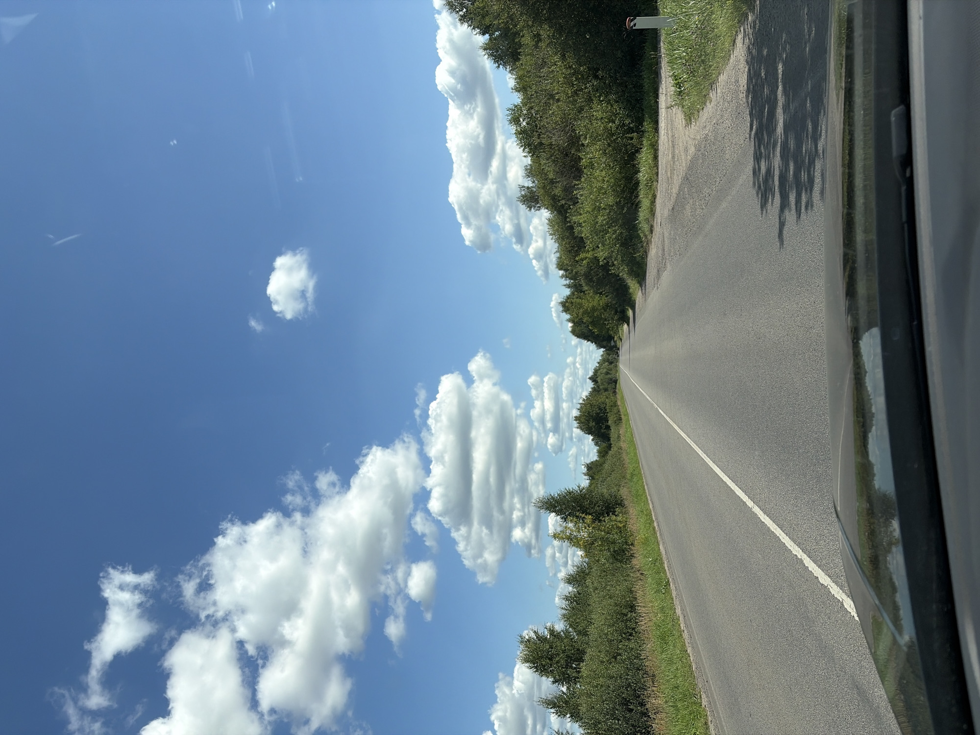 This is a vertical, eye-level, medium shot of a two-lane asphalt road stretching into the distance, lined by trees and vegetation on both sides. The sky is a vibrant blue, dotted with fluffy white cumulus clouds. The road has a white solid line on the left and a dashed line on the right, indicating it is a two-way street. The perspective is from inside a vehicle, with the hood of the car visible in the foreground. The scene appears to be in a rural area, likely on a sunny day. There are no people or animals visible in the image. The location is identified as Starye Psar'ki, Russia.
