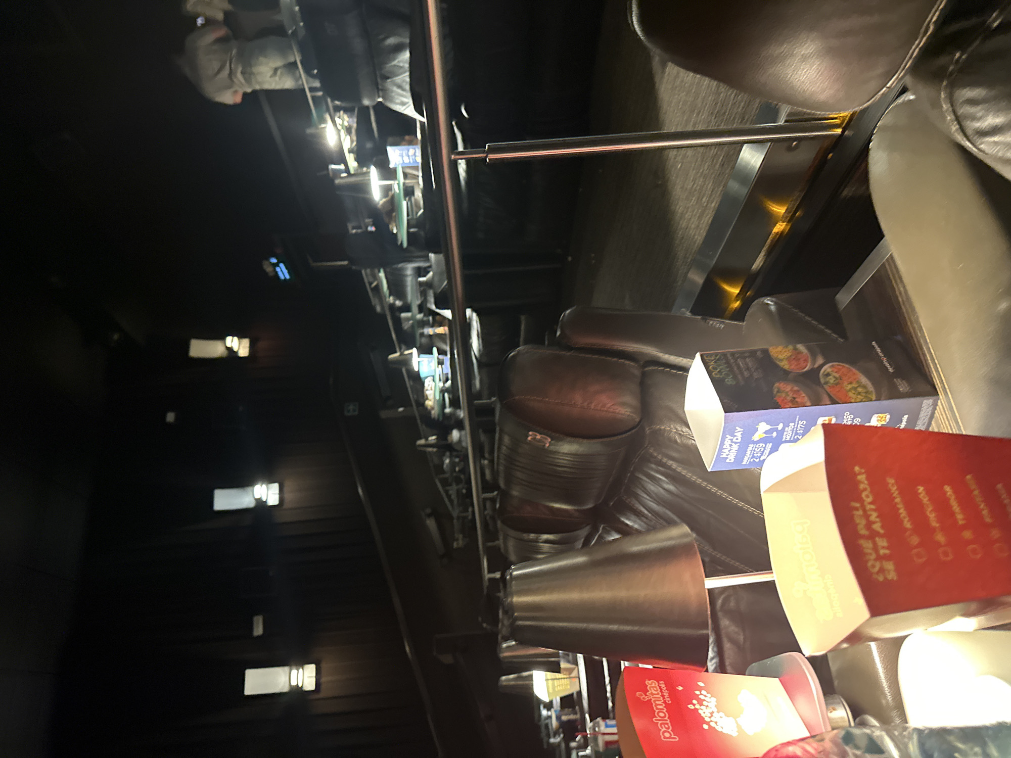 The image captures the dimly lit interior of a modern movie theater, likely a premium or VIP section, characterized by comfortable dark leather recliners and individual table-like surfaces. The perspective is from one of these seats, looking across the rows towards the front of the theater.
In the foreground, two empty or nearly empty movie snack containers are prominent. A red popcorn box clearly displays "palomitas cinépolis" (Cinépolis popcorn). Next to it, a white box asks "¿QUÉ PELI SE TE ANTOJA?" ("What movie do you fancy?") followed by genre options like "ROMANCE," "FICCIÓN" (fiction), and "TERROR." This box also advertises a "HAPPY DRINK DAY" with prices for "MARGARITAS" ($159) and "HORAS" ($175). A menu or advertisement for food items, possibly featuring bowls of rice or salads, is also partially visible on a table next to the seats.
A shiny silver metal railing runs horizontally through the midground, likely part of the seating arrangement or a shared table space. Small, soft lights emanate from built-in lamps or cup holders on the recliners, casting a warm glow. Further back, more rows of dark recliners are visible, with blurry figures of other moviegoers in the distance. The left side of the frame shows rectangular illuminated panels on a dark wall, possibly low-level lighting or exit indicators. The overall setting suggests a comfortable, evening entertainment experience, typical of a movie showing at a Cinépolis cinema in Huixquilucan de Degollado, Mexico, given the Spanish text and brand name.