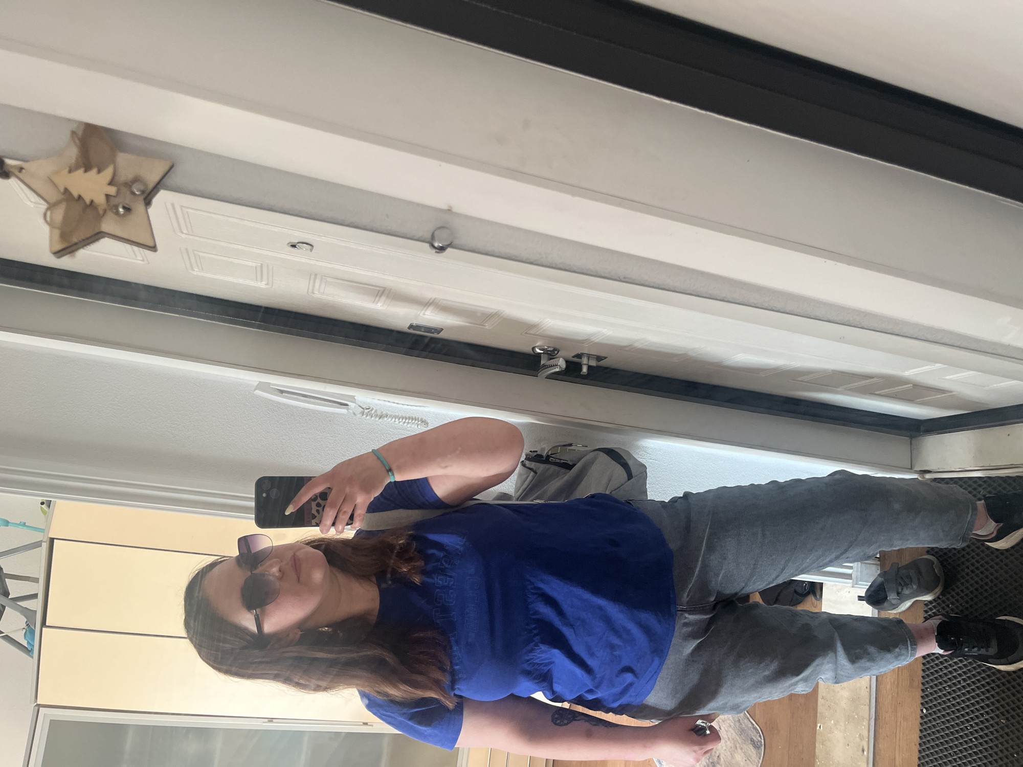 A woman is taking a mirror selfie in a dimly lit room. She is wearing a blue t-shirt, gray jeans, and sunglasses. She has brown hair and is wearing a turquoise bracelet on her left wrist. She is holding a smartphone in her right hand. In the background, there is a door with a star-shaped ornament hanging on it. There are also some cabinets and a mirror visible. The flooring is made of wood.