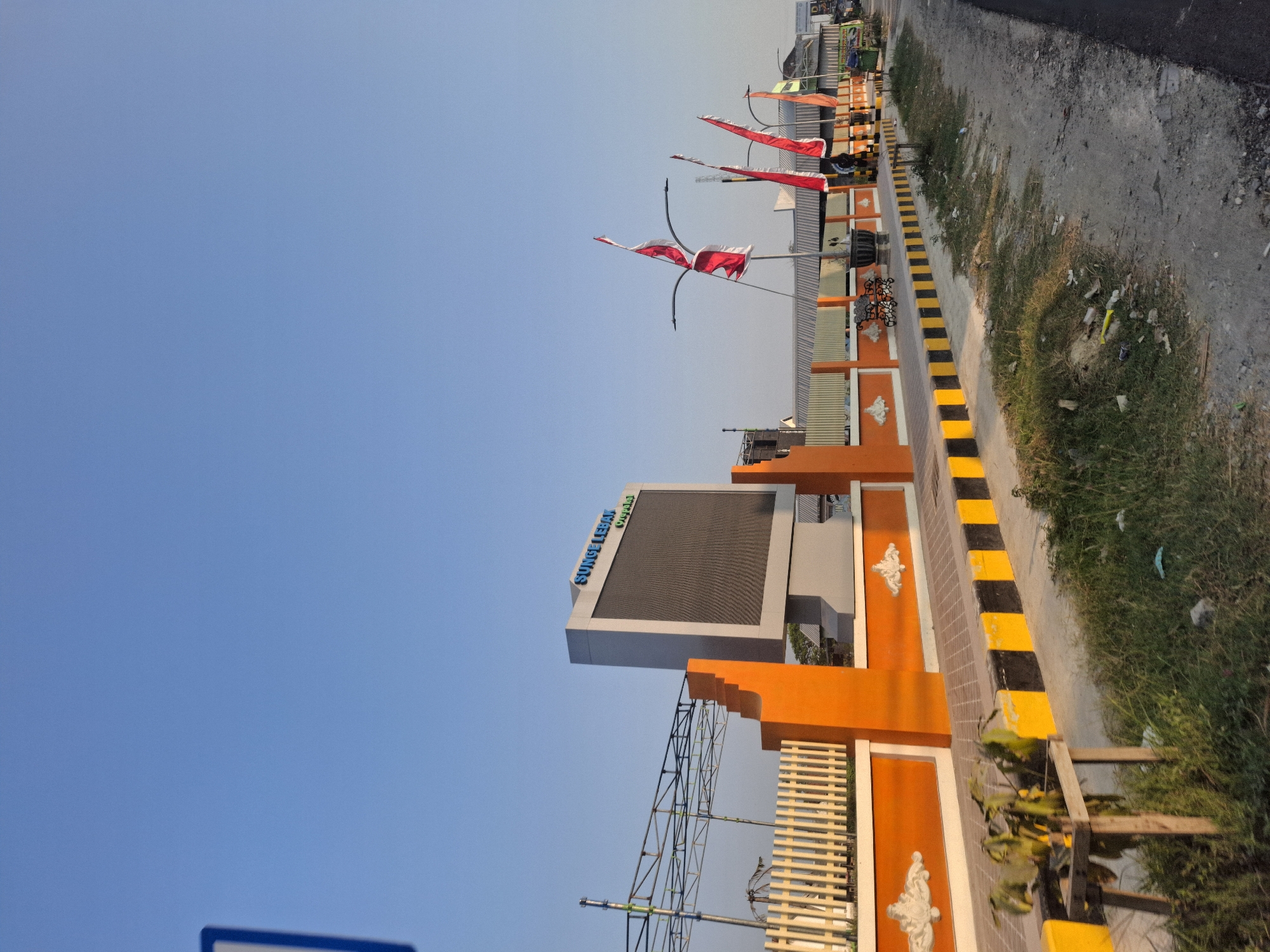 The image depicts an outdoor scene in Sungelebak, Indonesia. A large LED billboard with the text "Sungei Lebak" in blue lettering is prominently displayed. Adjacent to the billboard is an orange fence with decorative white accents. Several Indonesian flags, predominantly red and white, are flying on poles along the fence line. The scene includes a paved walkway with yellow and black striped borders, bordered by a grassy area and a dirt road. In the background, various structures and trees are visible, along with what appears to be a metal scaffolding structure. The sky is clear and blue, suggesting daytime. There are no discernible people or animals in the immediate foreground or midground.
