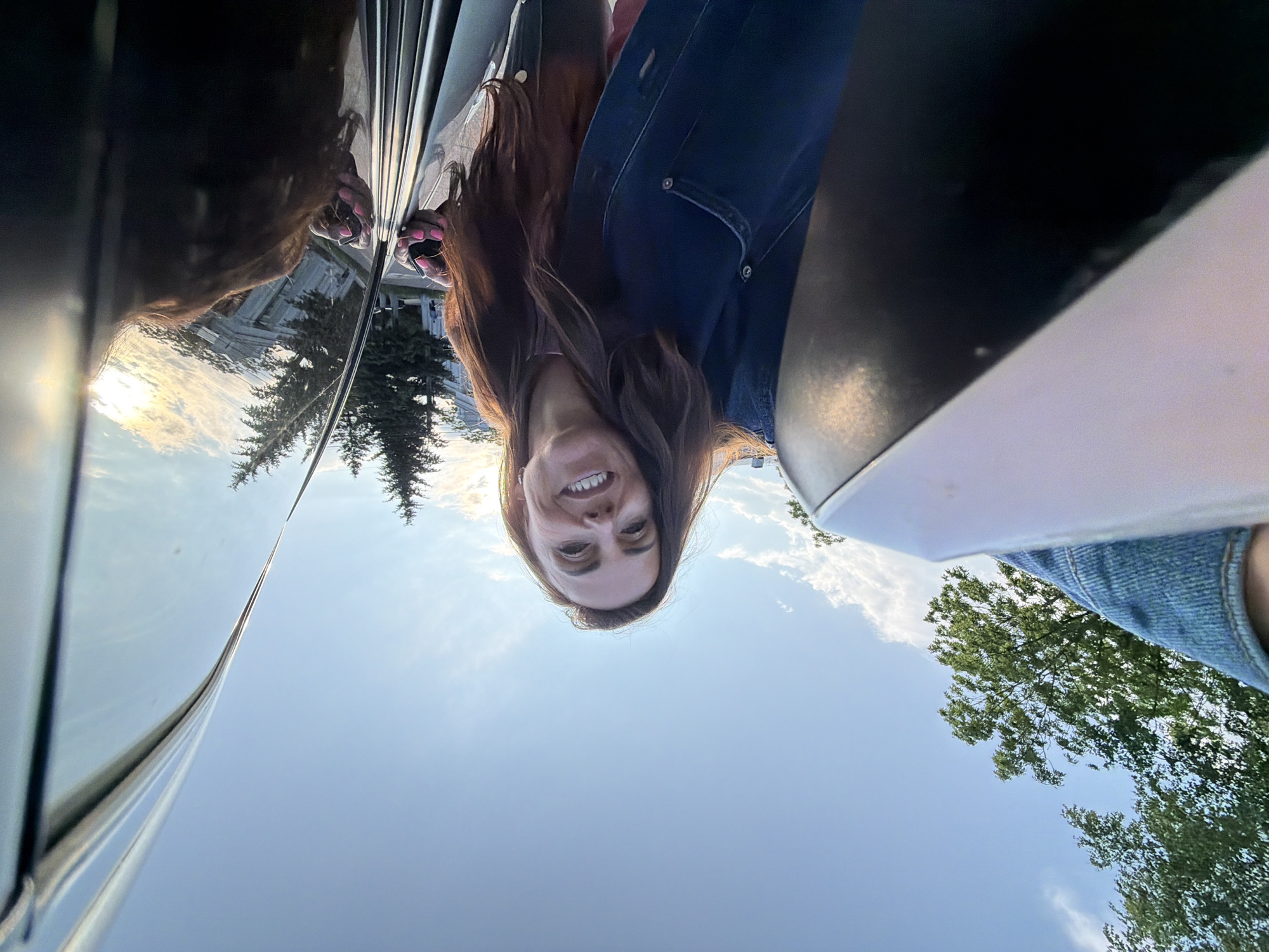 In Birobidzhan, Russia, a woman with long, dark hair and wearing a denim jacket smiles at the camera as she leans out of a car. Her right hand is visible, holding a set of keys. The car's shiny surface reflects the sky, trees, and buildings. The sky is blue with scattered clouds, and sunlight suggests it is either early morning or late afternoon. Trees and a building are visible in the background, with the overall scene appearing to be an outdoor setting within the city.
