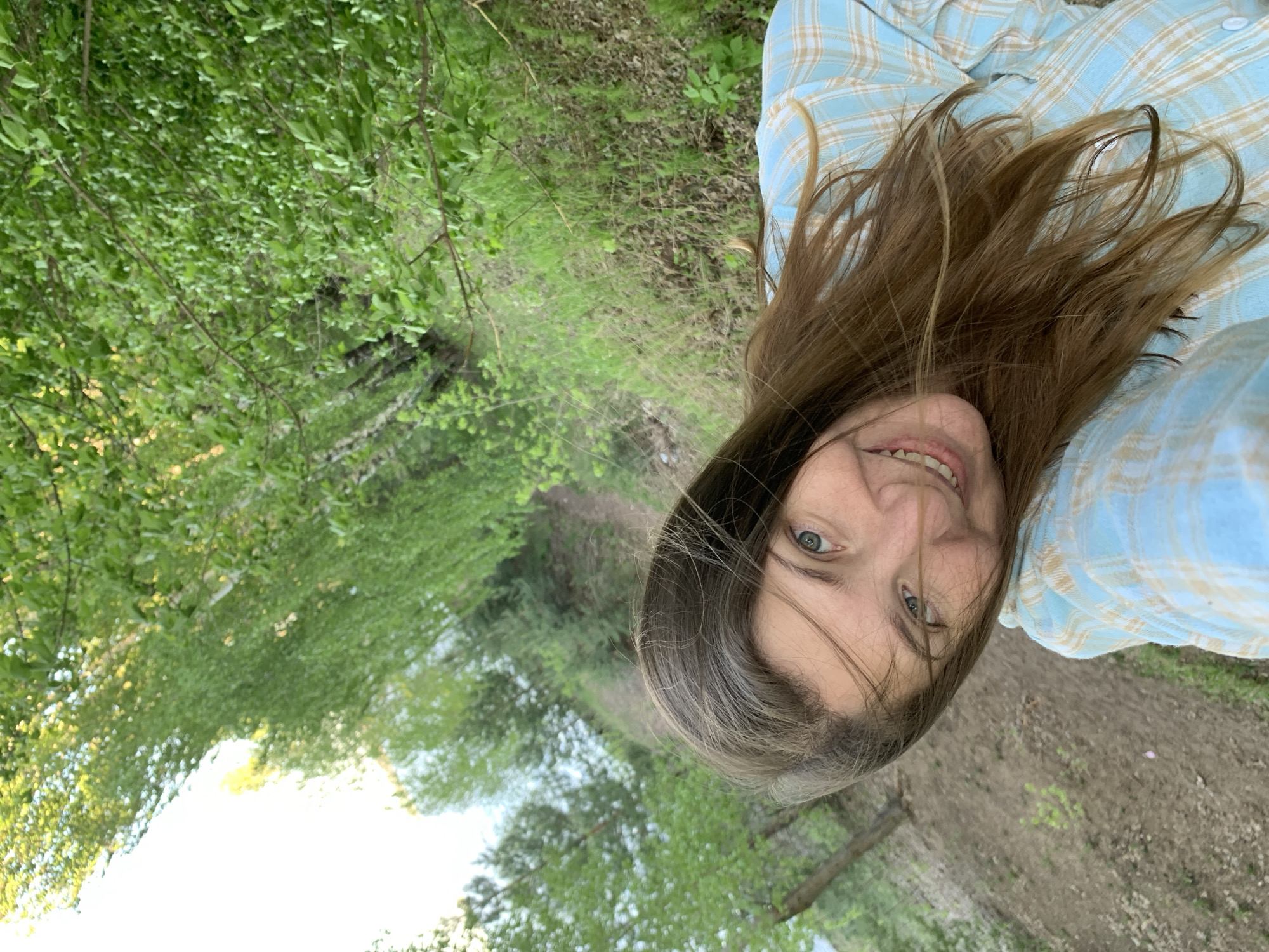 A woman with brown hair, wearing a blue and beige plaid shirt, is positioned in the foreground of a landscape. The background consists of a dirt path bordered by green vegetation and trees. The location appears to be outdoors, possibly a park or wooded area. The city is Chaikovsky, Russia.