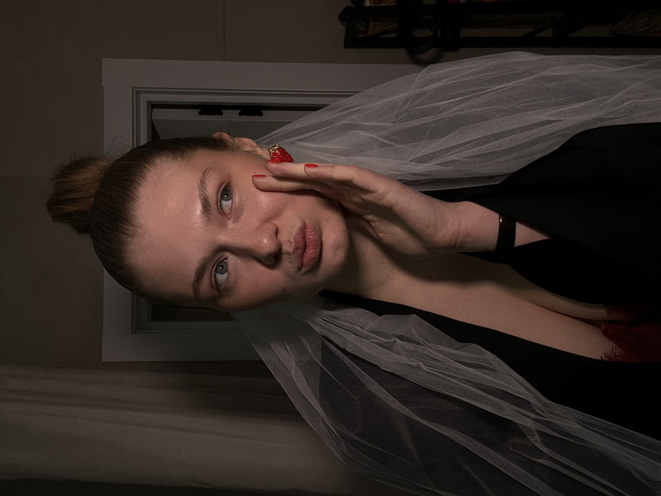 A female subject is captured indoors, positioned centrally within a dimly lit frame. She has fair skin, light-colored hair pulled into a high bun, and blue eyes looking to the viewer's right. Her lips are pursed in a pout. Her right hand (viewer's left) is raised, fingers splayed, with red nail polish, lightly touching her right cheek. A red, strawberry-shaped earring is visible on her right ear (viewer's left). She is draped in a translucent white fabric, resembling a veil, over a black garment with a deep V-neck that exposes a red, lace-trimmed undergarment. A dark-colored wristband or bracelet is present on her left wrist (viewer's right). The background features a light-colored curtain or drape to the viewer's left, a partially open white doorframe revealing a darker space to the viewer's right, and a dark, multi-shelf storage unit further right. The specific location within Kazan, Russia, cannot be determined from the visual evidence.