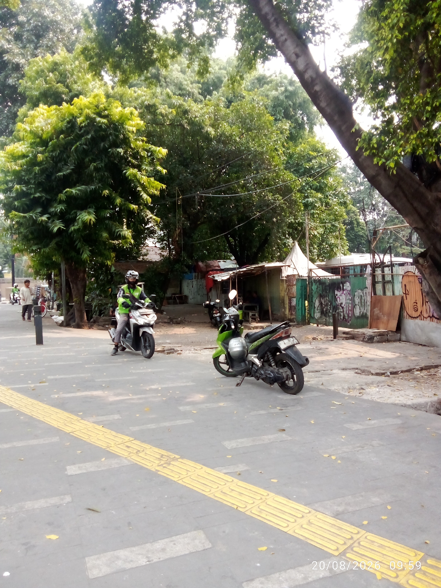 The image depicts a street scene in Central Jakarta, Indonesia, on August 20, 2026, at 09:59. In the foreground, a grey asphalt path is marked with a tactile paving strip of raised yellow squares, indicative of a pedestrian walkway designed for the visually impaired. Scattered fallen leaves litter the path.
Further into the scene, a person wearing a bright green jacket and helmet is riding a silver scooter towards the viewer. Another person, dressed in a light-colored shirt and dark pants, is walking away from the viewer on the same path. Behind them, to the left, another scooter is visible.
The background reveals a cluster of trees with dense green foliage, casting shadows on the path. Adjacent to the trees is a makeshift structure with corrugated metal walls, some of which are adorned with graffiti. A large tree trunk dominates the right side of the frame, partially obscuring the sky. The overall environment suggests a public urban space, likely a sidewalk or pedestrian area alongside a road, with elements of both natural and constructed infrastructure.