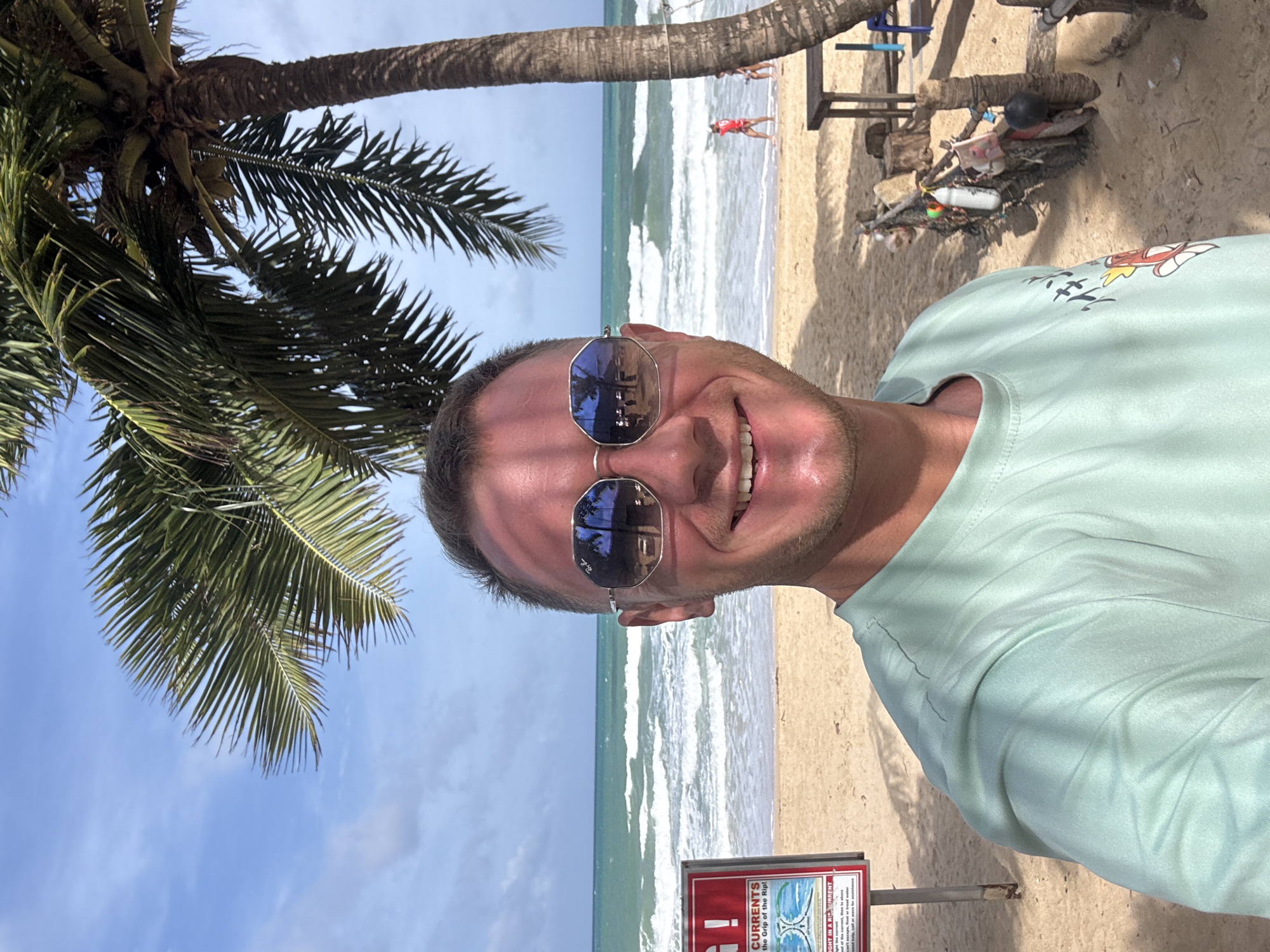 A man with short brown hair and sunglasses is taking a selfie on a beach in Rawai, Thailand. He is wearing a light green t-shirt with a graphic print on the left side. Behind him, the ocean has large waves, and palm trees hang overhead. A red and white sign warning about rip currents is visible on the left side of the image, and a wooden table and chairs are on the right. The sky is blue with some clouds.