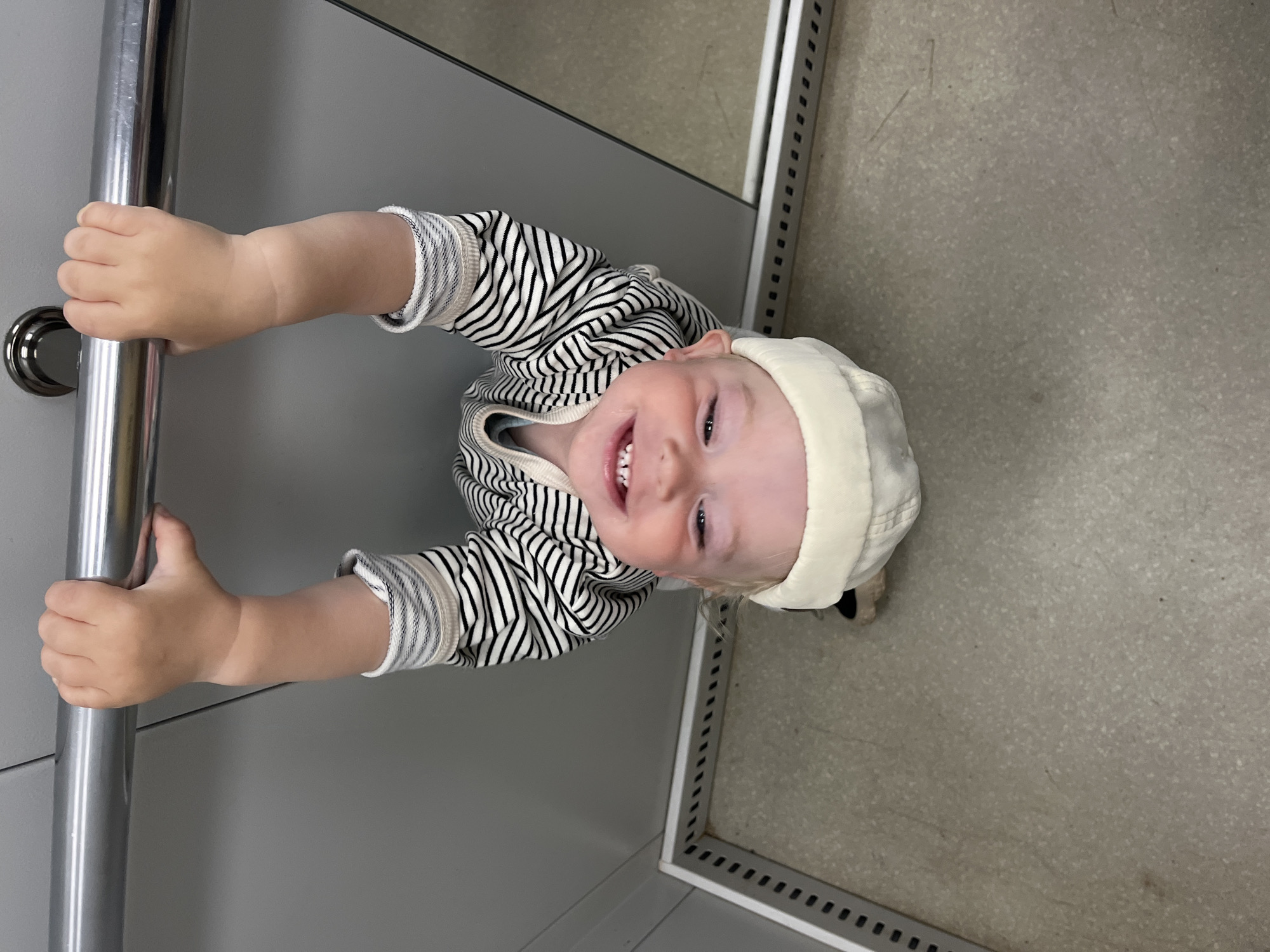 A toddler hangs upside down from a metal handrail, grasping it with both hands. The child wears a striped long-sleeved shirt and a cream-colored hat. The background consists of grey walls and a speckled floor. The image is taken in an indoor environment, possibly an elevator or a public space with handrails. The child is smiling. There is no information in the image to determine the location in the city of Chaikovsky, Russia.