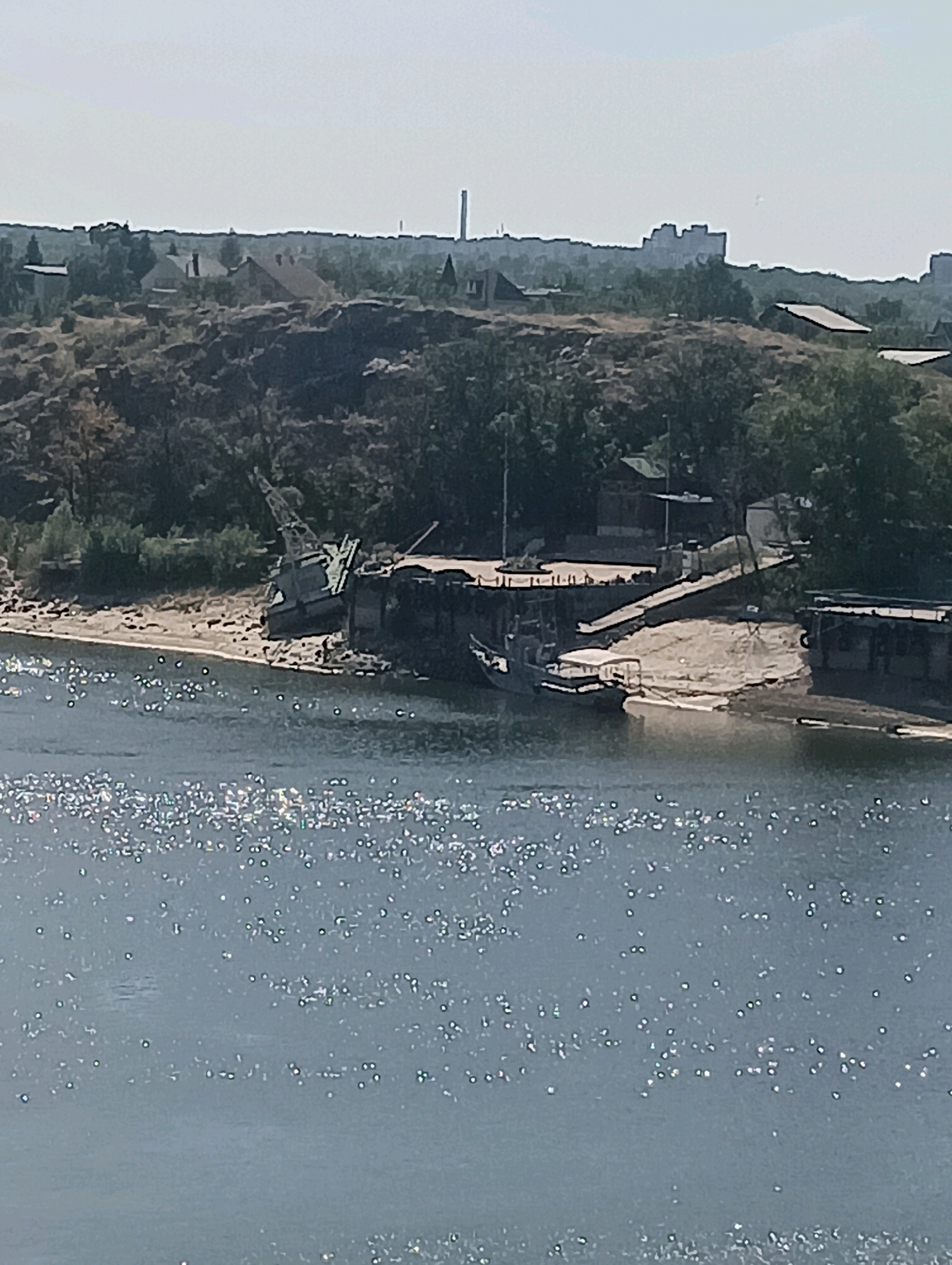 In this aerial view of a body of water and a shoreline, the water in the foreground is illuminated by bright sunlight, creating a shimmering effect with numerous reflections. The shoreline, which is a mix of sand and rocky terrain, features several objects. Prominently, a large, industrial-style crane is situated on the shore, appearing partially submerged or very close to the water's edge. Next to the crane, there is a concrete structure that resembles a dock or a pier. A small boat with a white canopy is visible near the pier. Further inland, a collection of buildings, including houses and industrial structures, are nestled amongst trees on a gentle slope. In the background, the silhouette of a city skyline is visible, characterized by several buildings and a tall smokestack. The sky is clear and bright.