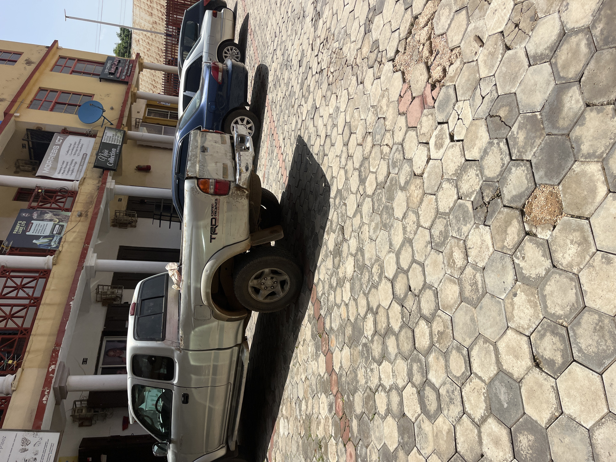 The image shows a street scene in Gura Top, Nigeria, with several parked cars and a building. A silver Toyota pickup truck is prominently featured in the foreground, with "TRD OFF ROAD" visible on its side. Behind it, a blue car and a silver sedan are parked. The building on the left has a yellow facade with red-trimmed windows and several signs. One sign reads "MORREBS ICT" and lists services like "Business Centre," "Coding School," and "Graphics Design." Another sign says "Nique Collection." The ground is paved with hexagonal concrete tiles, some of which are cracked and worn. The lighting suggests it is daytime, possibly mid-morning or afternoon, with clear skies. The scene appears to be a typical urban street with commercial establishments. No people are visible in the frame.