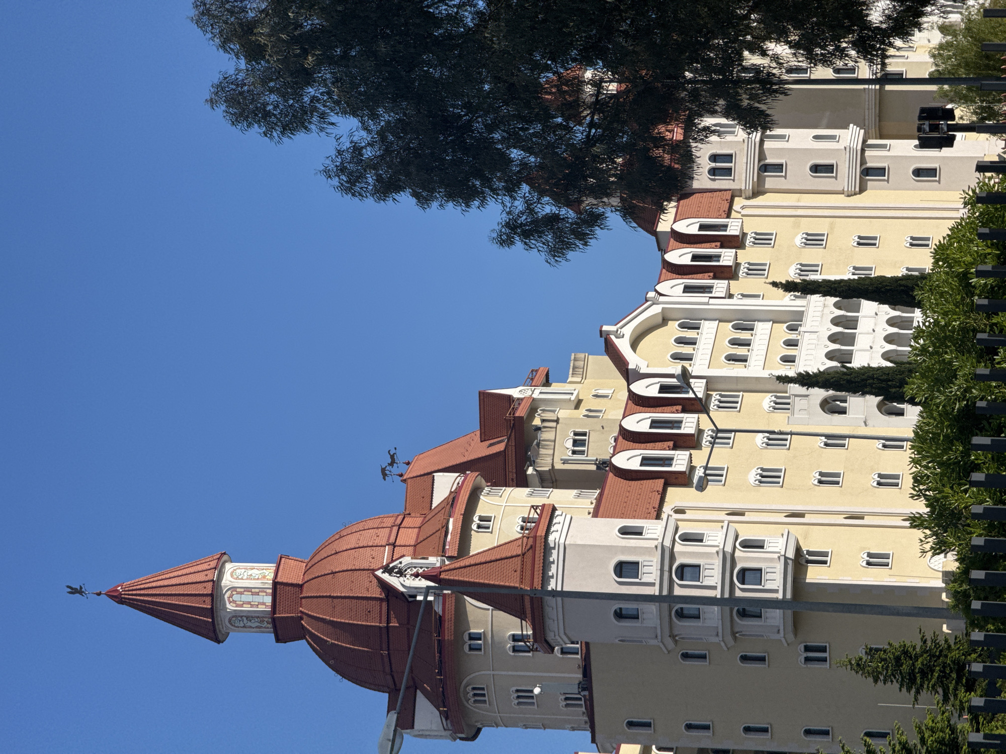 The image shows a multi-story building in Sirius, Russia, characterized by a cream-colored facade and red tiled roofs. The architectural style features prominent arches and decorative window frames. The building's structure includes a tower topped with a pointed spire, adorned with a weather vane. Clear blue sky occupies a significant portion of the frame. The building is partially framed by foliage, including a large tree with dark green leaves at the top, and other greenery at the sides, suggesting a park or garden setting. A portion of a street light pole is visible on the left side.