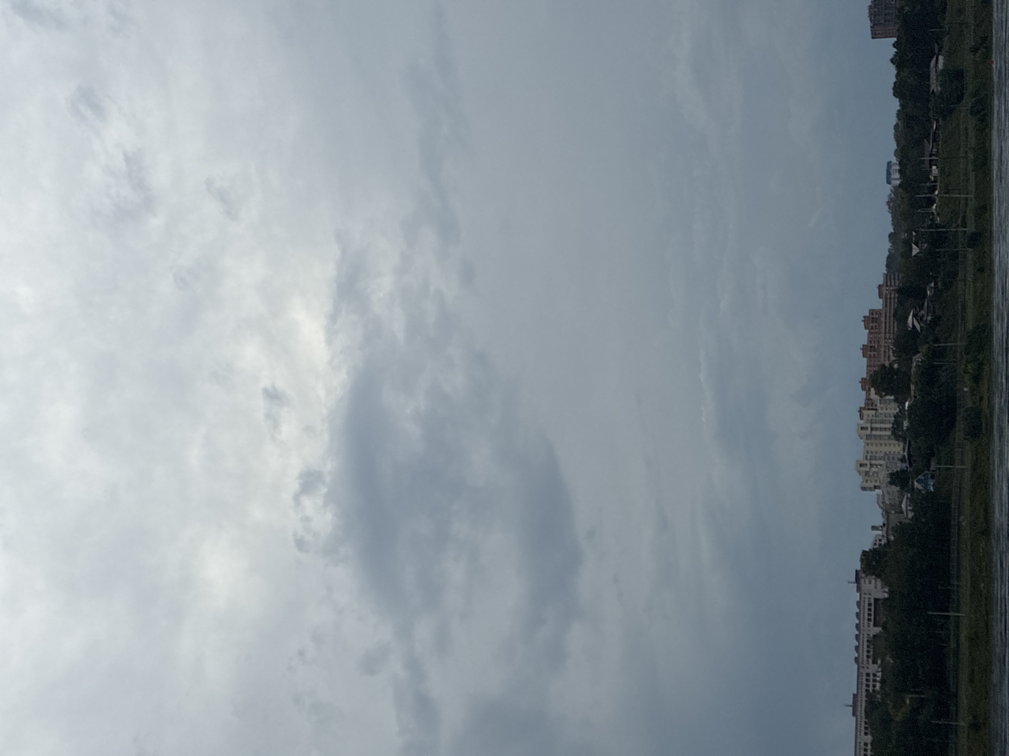 The image depicts a cityscape under a cloudy sky. In the foreground, a body of water, likely the Angara River, is visible, with ripples on its surface. Beyond the water, a line of trees separates the riverbank from the city. The city itself features a mix of buildings, including several multi-story apartment complexes and smaller houses. The sky is overcast with layers of gray clouds, obscuring the sun and giving the scene a somber or potentially stormy atmosphere. The buildings appear to be located on a gentle slope leading down towards the river. The overall impression is that of a residential area within a city, viewed from across a river on a cloudy day. Given the context, this scene is likely located in Irkutsk, Russia, along the Angara River. There are no people or visible actions taking place.