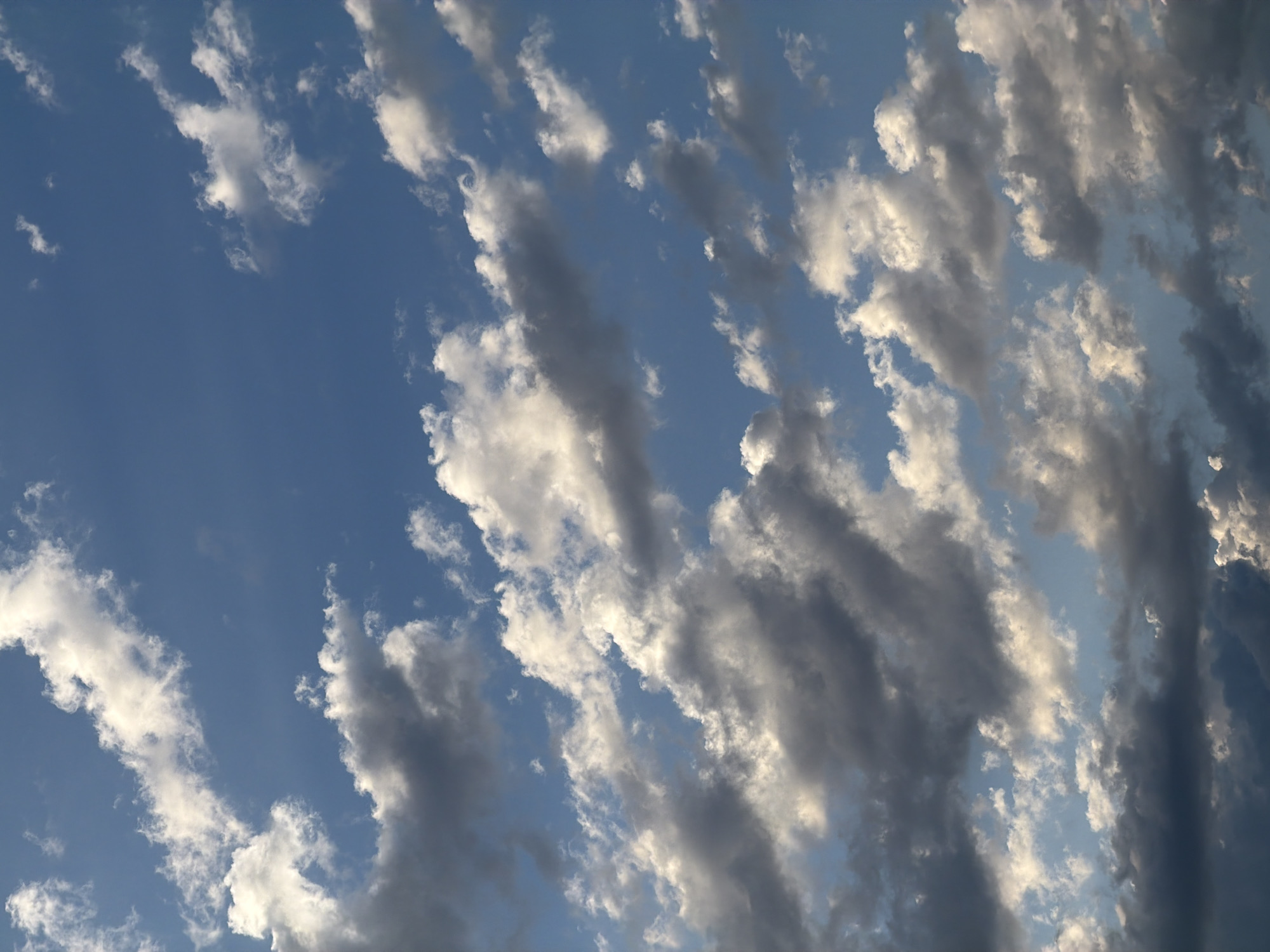 The image shows a sky filled with a mix of white and gray clouds against a bright blue sky. Some clouds are wispy and translucent, catching the sunlight, while others are darker and more substantial, suggesting depth. The overall impression is of a dynamic sky with varying cloud formations. The image does not depict any urban elements, people, or specific activities, and therefore cannot be localized to Arcahueja, Spain, or any other city.