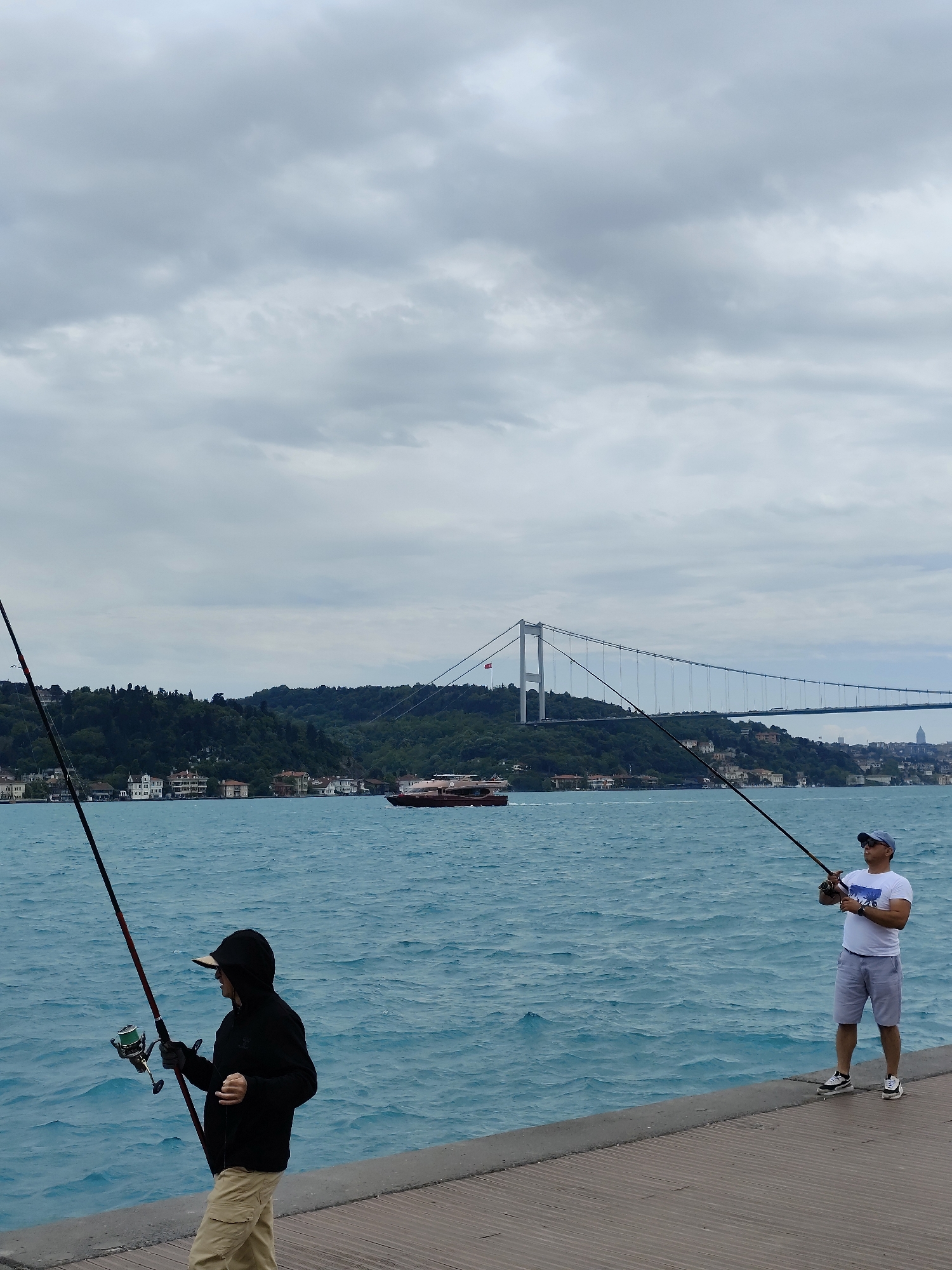 Two individuals are fishing on a pier in Sarıyer, Turkey. A young person in a black hooded jacket and tan pants stands on the left, holding a fishing rod. An adult male in a white T-shirt, gray shorts, and a baseball cap stands on the right, also holding a fishing rod. Between them is the Bosphorus, with a passenger ferry traversing the water and houses on the shoreline. In the background, the Bosphorus Bridge connects the European and Asian sides of Istanbul. The sky is overcast with gray clouds. The pier appears to be made of wood or a wood-like material.