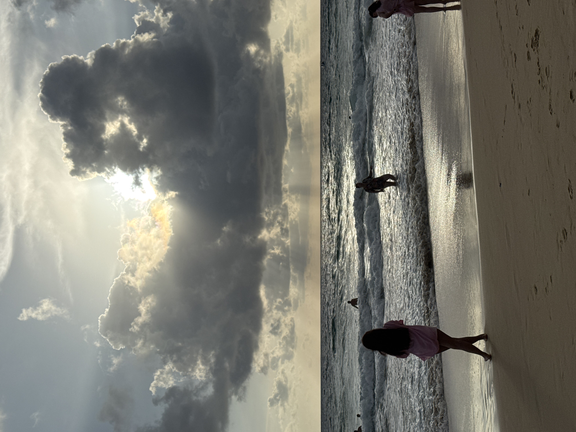 The image depicts a beach scene at Karon, Thailand, during sunset. The sky is dramatic with heavy, dark clouds partially obscuring the sun, which casts rays of light through gaps. The ocean has moderately sized waves with whitecaps, and there are several people in the water and on the shore.
In the foreground, on the sandy beach, a person wearing a pink cover-up stands with their back to the camera, facing the ocean. To the right, another person, also in pink, is partially visible at the edge of the water. Further out in the ocean, a person in darker attire is standing in the waves, and other figures can be seen further offshore. The wet sand reflects the light from the sky. The overall atmosphere is serene and a bit moody due to the cloud cover and the setting sun.