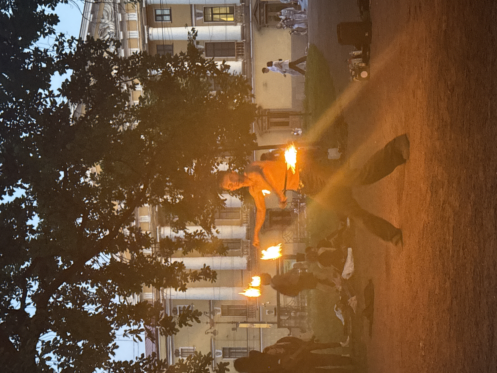 The image depicts an outdoor evening scene in the historic center of Saint Petersburg, Russia. The primary subject is a shirtless male performer, positioned centrally, engaged in a fire show. He actively swings two flaming props, one in each hand, producing distinct lens flares across the foreground. He wears dark trousers and light-colored shoes. To his left, another individual, largely silhouetted, also displays flames above their head, suggesting a secondary performer.
In the mid-ground and background, approximately fifteen other individuals are present. Several people are seated or lying on the reddish-brown dirt/gravel ground, seemingly observing the performance. On the right side, a couple walks away from the camera on a paved path, and further back, a small group including a child and a dog is visible.
The background features multi-storied buildings characteristic of Saint Petersburg's neoclassical or Empire architectural style. These structures display cream or yellow facades, white classical columns, rectangular windows, and ornate pediments with sculptural reliefs and friezes. Mature deciduous trees with dense, dark foliage are silhouetted against a pale blue twilight sky, framing the upper portion of the scene.
On the far right in the foreground, performance equipment is visible on the ground, including a dark cylindrical object (possibly a speaker or container) and a white box or cooler, alongside other smaller items. The scene is illuminated by the prominent flames and residual ambient light from the early evening sky.