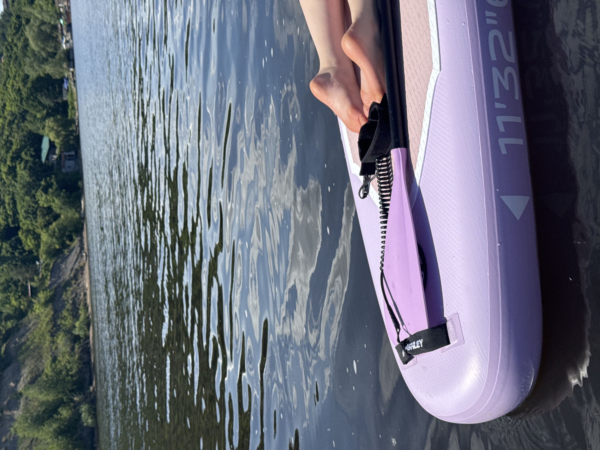 The image presents a peaceful outdoor scene on a sunny day, likely in Усть-Курдюм, Russia. The viewpoint is slightly elevated, looking down at a stand-up paddleboard (SUP) and across a body of water towards a distant, tree-lined shore.
The main subjects are the bare feet and lower legs of a person, resting comfortably on a light lavender or purple stand-up paddleboard. The person's stance suggests relaxation, possibly sitting or kneeling on the board.
The primary object is the brightly colored paddleboard, which has a textured surface where the feet are placed. A black coiled leash or tether is visible, attached to the board near the feet, and features a strap with the word "ASHLEY" printed on it. Further along the side of the paddleboard, the text "11'32''" is clearly legible, likely denoting the board's dimensions. The water around the board exhibits gentle ripples, reflecting the clear blue sky and scattered white clouds, indicating calm conditions.
In the background, a verdant shoreline extends across the frame, densely covered with green trees. Some faint structures or buildings can be seen nestled within the foliage, suggesting a natural area bordering human settlement.
The activity taking place is recreational, with the individual enjoying the water on the paddleboard. The bright sunlight and reflections on the water confirm it is daytime, likely midday or early afternoon. The scene evokes a sense of tranquility and leisure, emphasizing enjoyment of nature and warm weather.