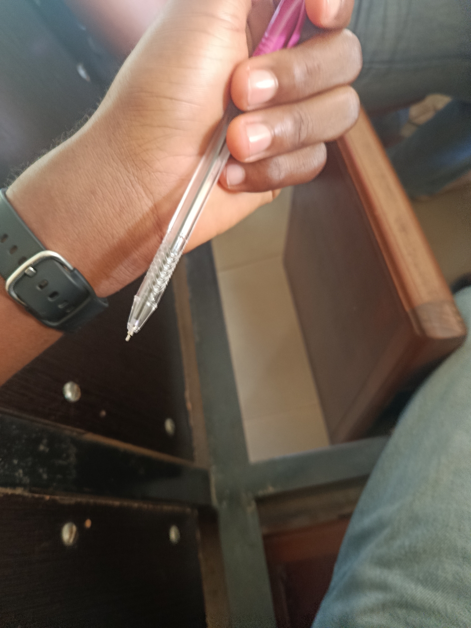 A close-up, vertical shot shows a person's right hand holding a pink and clear ballpoint pen. The pen is positioned diagonally from the top right to the bottom left of the frame, with its tip pointing downward. The hand is dark-skinned, and the person is wearing a black watch with a silver buckle on their left wrist. The background consists of a wooden surface, possibly a desk or chair, and blurred figures in denim clothing. The lighting is dim, with some glare on the watch and the pen. The scene appears to be indoors, possibly in a classroom or office setting.