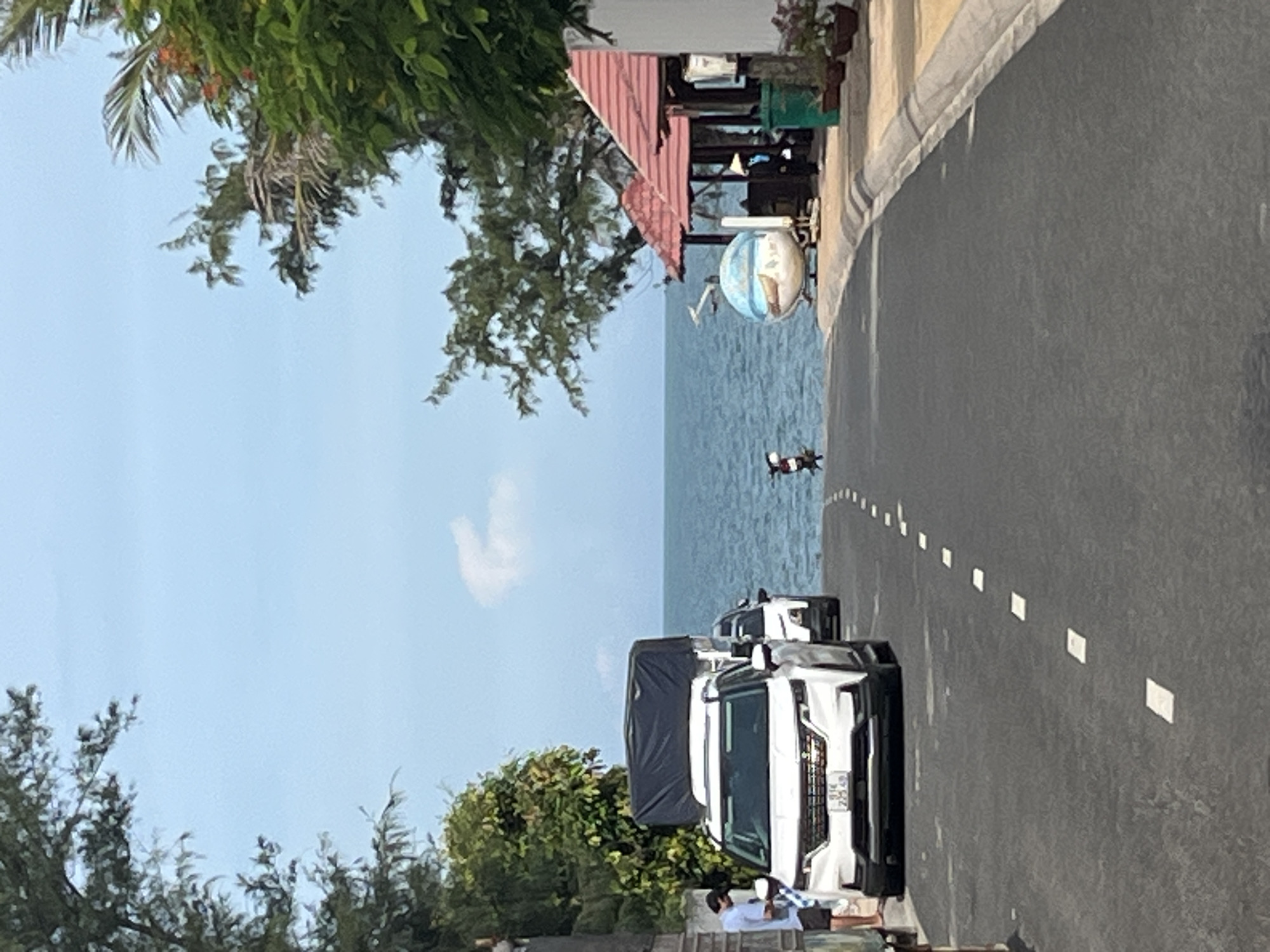 The image depicts a street in Mũi Né, Vietnam, leading towards the ocean. The road is paved with asphalt and has white dashed lines indicating lanes. On the left side of the road, a white SUV is parked, and a man in a blue shirt is standing beside it, looking at his phone. Further down the road, another white SUV is visible, followed by a covered trailer. Trees and foliage line the left side of the road.
On the right side of the road, there are buildings with red roofs, possibly shops or restaurants, and a small spherical decorative object with a beach scene painted on it. A person wearing a conical hat is standing near one of the buildings. The ocean is visible in the background, with a clear blue sky above. A small motorbike is parked on the side of the road in the distance. The overall scene suggests a coastal town or resort area.