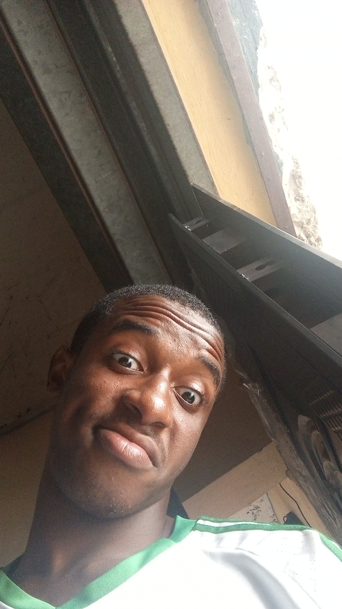 A young man is captured in a close-up selfie, taken indoors from a low, upward-tilted angle. He gazes directly into the camera with a wide-eyed, expressive look and slightly pursed lips, suggesting a surprised or playful emotion. His eyebrows are furrowed, creating horizontal lines on his forehead. He has dark skin and short, dark, curly hair. He is wearing a white shirt or jersey featuring green trim around the collar and a green stripe on the shoulder.
The background is out of focus and highly angled. It reveals abstract structural elements, including dark, parallel beams or panels on the left and top. Towards the upper right, a lighter, possibly cream or yellow-colored surface is visible, adjacent to a rough, textured area that appears to be a white or light grey wall or concrete. Bright light illuminates the scene, particularly from the right, suggesting a strong light source like a window or artificial lighting. No text is visible in the image.