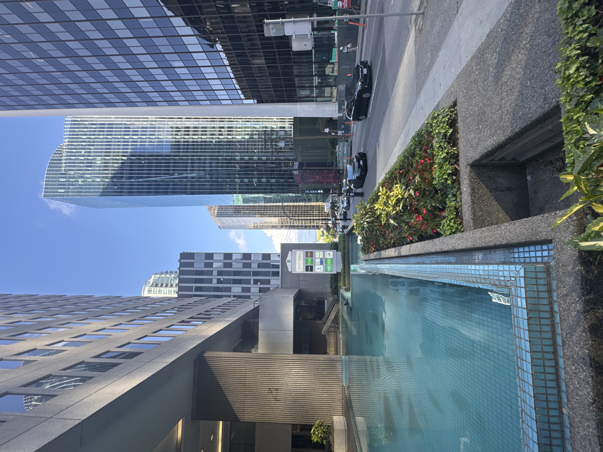 The image shows a cityscape in Vancouver, Canada, notable for its modern architecture and urban design elements. A long, rectangular, aquamarine tiled pool dominates the foreground. Adjacent to the pool is a raised planter filled with greenery. The surrounding architecture includes modern office buildings with glass curtain walls that reflect the sky. Cars are parked on the street, and there's a sign with multiple logos visible in the background.