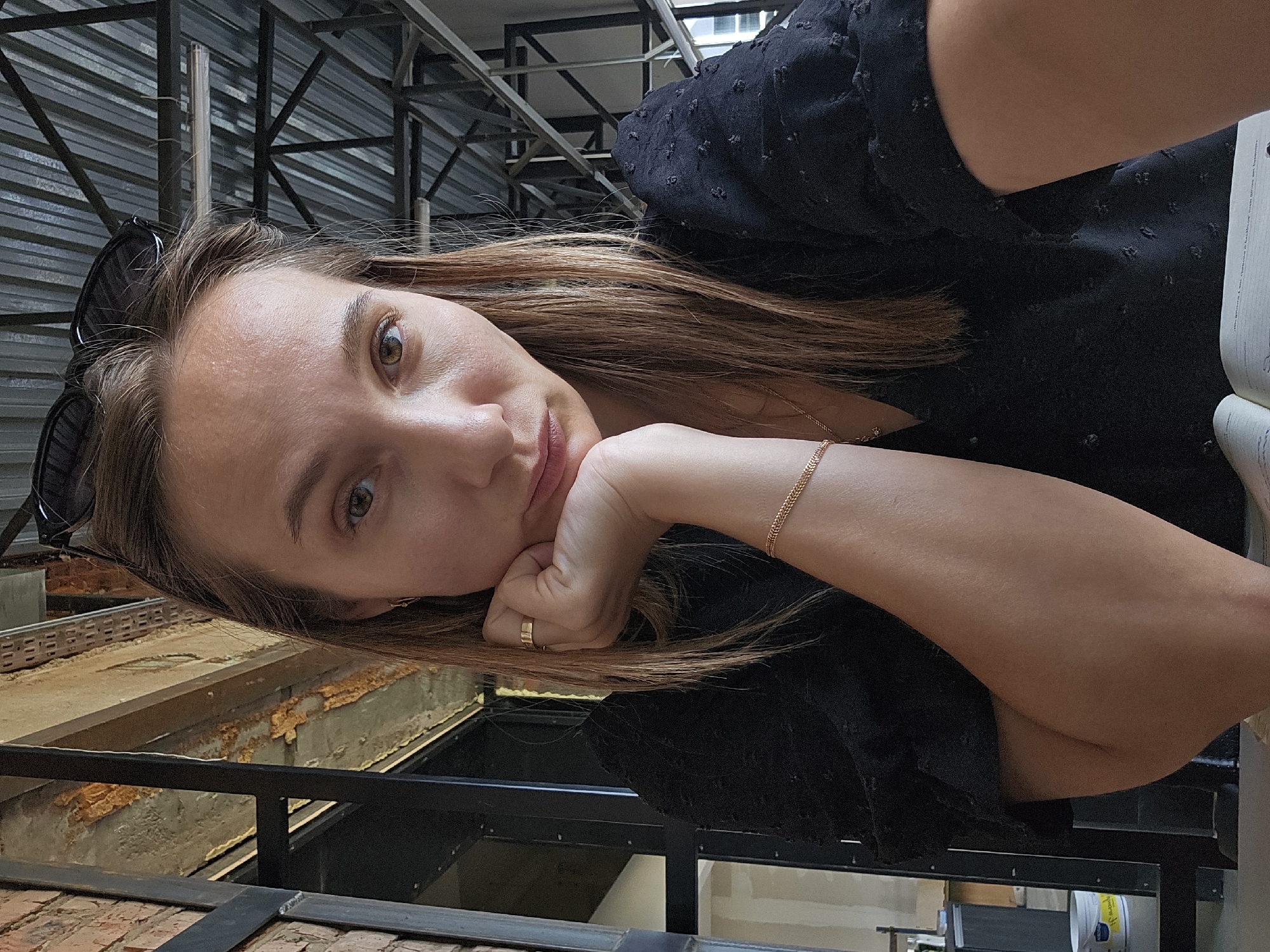 A woman with brown hair is in the city of Chaikovsky, Russia. She is propping her head on her hand, wearing a gold ring and a gold bracelet. Sunglasses are resting on her head. She is wearing a black textured shirt. The location is an industrial setting with metal beams and a partially constructed area. There's a brick structure with metal framing visible.