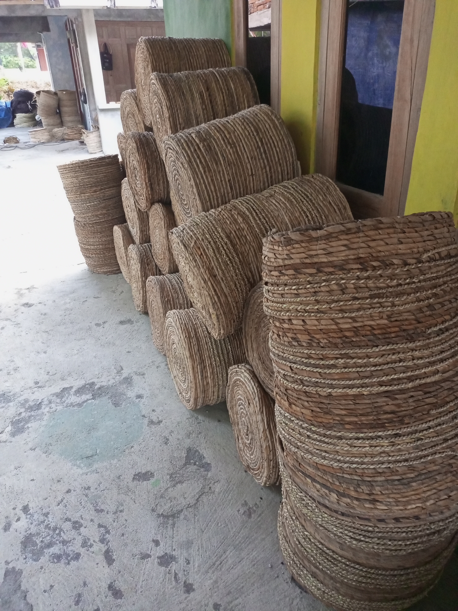 In Nanggulan, Indonesia, a collection of woven baskets is displayed on a concrete floor. The baskets, made from natural fibers, are stacked in various sizes and orientations. Some are cylindrical and tall, while others are flatter and round. The foreground features a large, wide basket with intricate weaving. Behind it, a diagonal row of progressively smaller baskets is visible, creating a sense of depth. In the background, more baskets are stacked along a wall, some appearing to be partially obscured by other objects and structures. The environment suggests an outdoor or semi-outdoor workspace, possibly a workshop or market stall. The lighting is natural, casting soft shadows on the ground. The overall impression is one of artisanal craftsmanship and abundance.