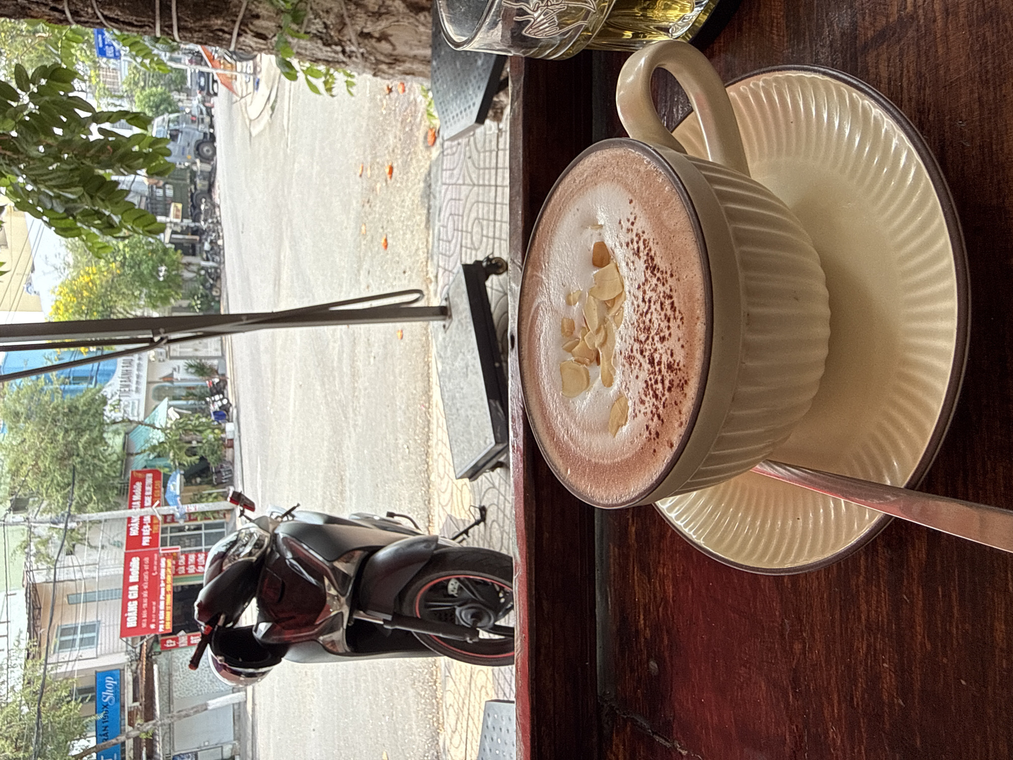 The image captures a street scene in Nam Nha Trang Ward, Vietnam, viewed from an indoor or semi-outdoor seating area, likely a cafe. In the immediate foreground, a light-colored ceramic cup with a handle holds a frothy beverage, possibly hot chocolate or coffee, topped with sprinkled powder and slivered almonds. The cup rests on a matching ribbed saucer, which also holds a metal spoon. Part of a glass with a yellowish liquid is visible further back on the dark wooden table.
The background unfolds into a vibrant street view. A dark-colored motorbike, possibly a scooter, is prominently parked on the sidewalk with a helmet resting on its seat. Further down the street, other vehicles, including a grey truck, can be seen. Buildings of various sizes line the street, many featuring shopfronts. Green trees and foliage are scattered throughout the scene, with a thick tree branch partially framing the top left.
Visible Vietnamese text on the shop signs includes:
- A red sign: "HOÀNG GIA MOBILE," "Mở bán iPhone Mới" (Selling new iPhones), "Phụ kiện" (Accessories), and "SỬA CHỮA ĐIỆN THOẠI" (Phone Repair).
- Another sign with "TRẦN 198x Shop."
The scene is set during the daytime, under what appears to be clear weather, with natural light illuminating the street. The overall mood suggests a relaxed observation of daily life in the Vietnamese street environment. No individuals are distinctly visible in the frame.