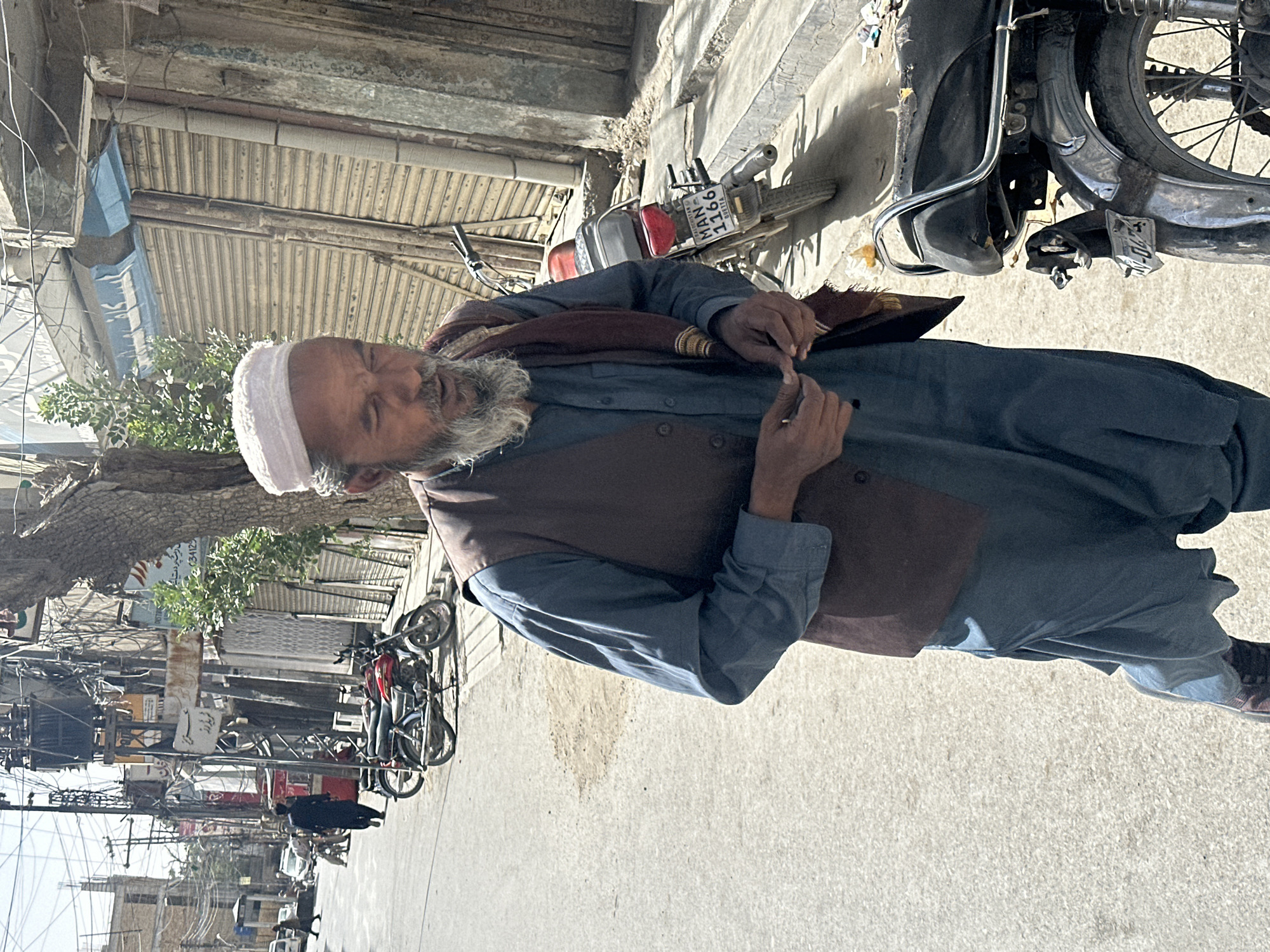 The image depicts a man wearing a white skullcap, a dark vest, and a blue shalwar kameez, walking in a street in Quetta City Tehsil, Pakistan. He has a gray beard and appears to be looking down, holding something small in his hands. In the background, there are several motorcycles parked along the side of the street. Buildings with closed shop shutters line the street, and there are numerous electrical wires and poles overhead, indicative of a bustling urban environment. The street surface is paved with asphalt. The lighting suggests it is daytime, with strong sunlight casting shadows.