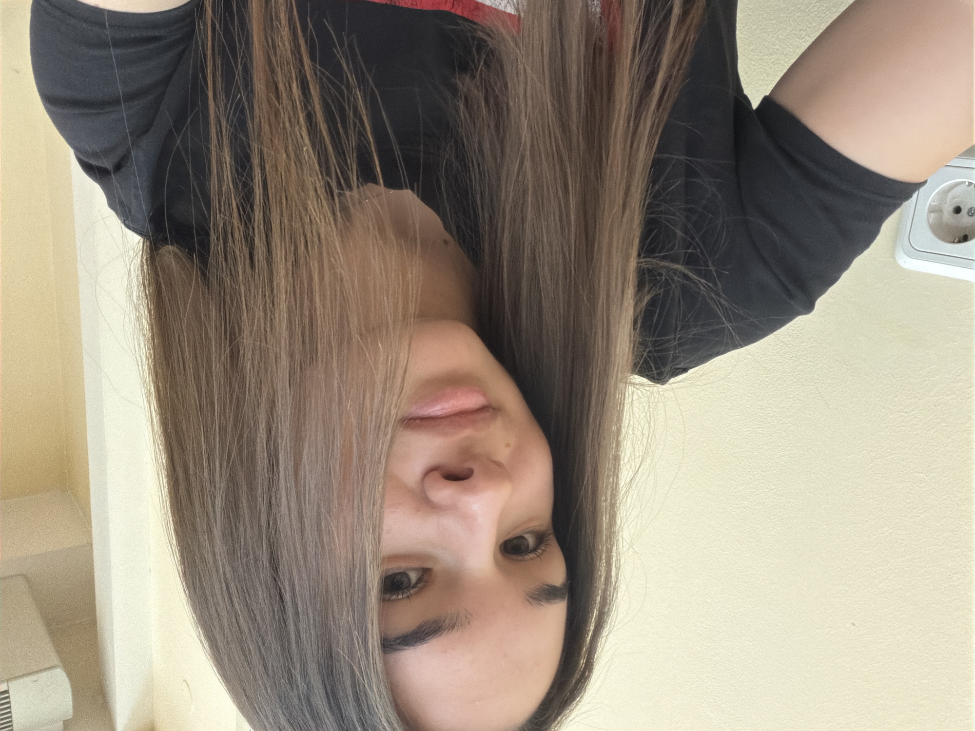 A woman with long, light brown hair is facing the camera. She is wearing a black t-shirt. The background consists of a light yellow wall, a white shelf unit, and a European-style electrical outlet. The location is presumed to be indoors. No identifying elements suggest the city of Sofia, Bulgaria.