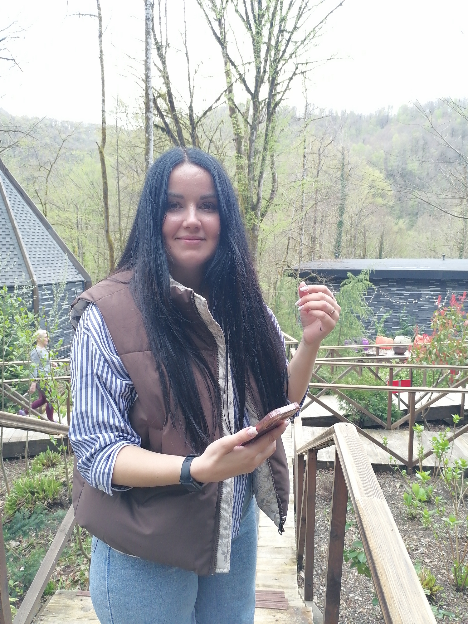 A young woman is depicted in a medium shot, smiling gently at the camera. She has long, dark, straight hair with a center part. She is wearing a brown puffer vest over a blue and white striped long-sleeved shirt and light blue jeans. On her left wrist, she wears a black digital watch, and a delicate ring is visible on the ring finger of her raised left hand, which has white-tipped nails. Her right hand holds a brown smartphone.
The scene appears to be an outdoor setting, possibly a resort or nature park. In the background, lush green forested hills and mountains are visible under an overcast bright sky, indicating daytime. Numerous trees, some with new spring foliage and others with bare branches, populate the landscape. To the left, a modern building with a steeply pitched, dark shingled roof can be seen. To the right, another dark, rectangular building with textured siding is visible, surrounded by colorful potted plants.
The woman stands on a wooden walkway or deck with matching railings. Another person, seen in the mid-ground to the left, is walking away from the camera on a wooden path, wearing a light-colored top and dark trousers. The overall environment suggests a blend of natural beauty with contemporary architectural elements. No visible text is discernible in the image.