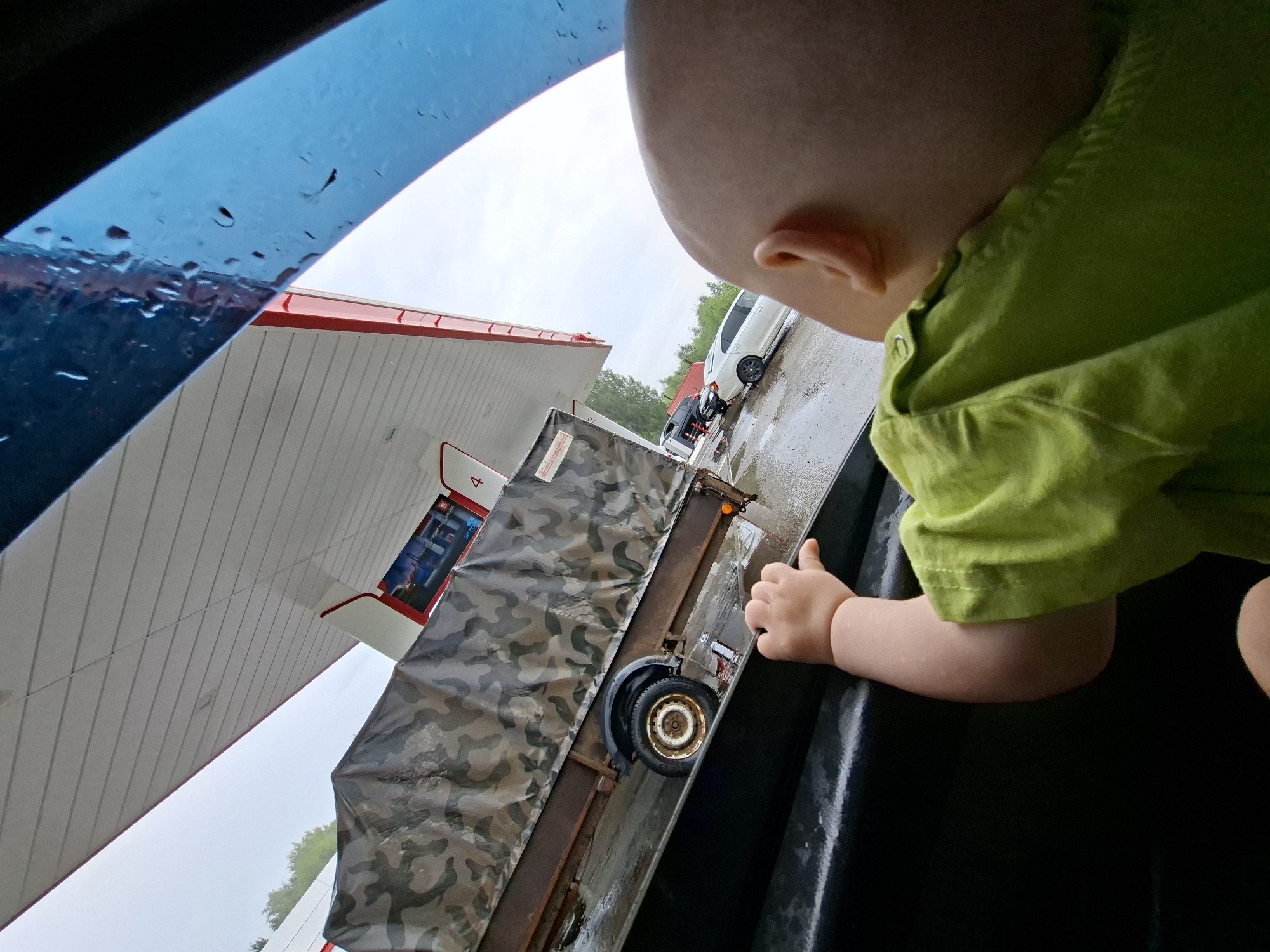 A baby wearing a lime green shirt leans out of a vehicle window, looking towards a gas station. The baby's left hand rests on the car's interior edge. Outside, a trailer covered in camouflage tarp is parked under the gas station's canopy, which is white with red trim. The ground is wet, indicating rain, and a white car is visible to the right of the trailer. The sky is overcast. The scene appears to be at a gas station located in the city of Krilovo, Russia.